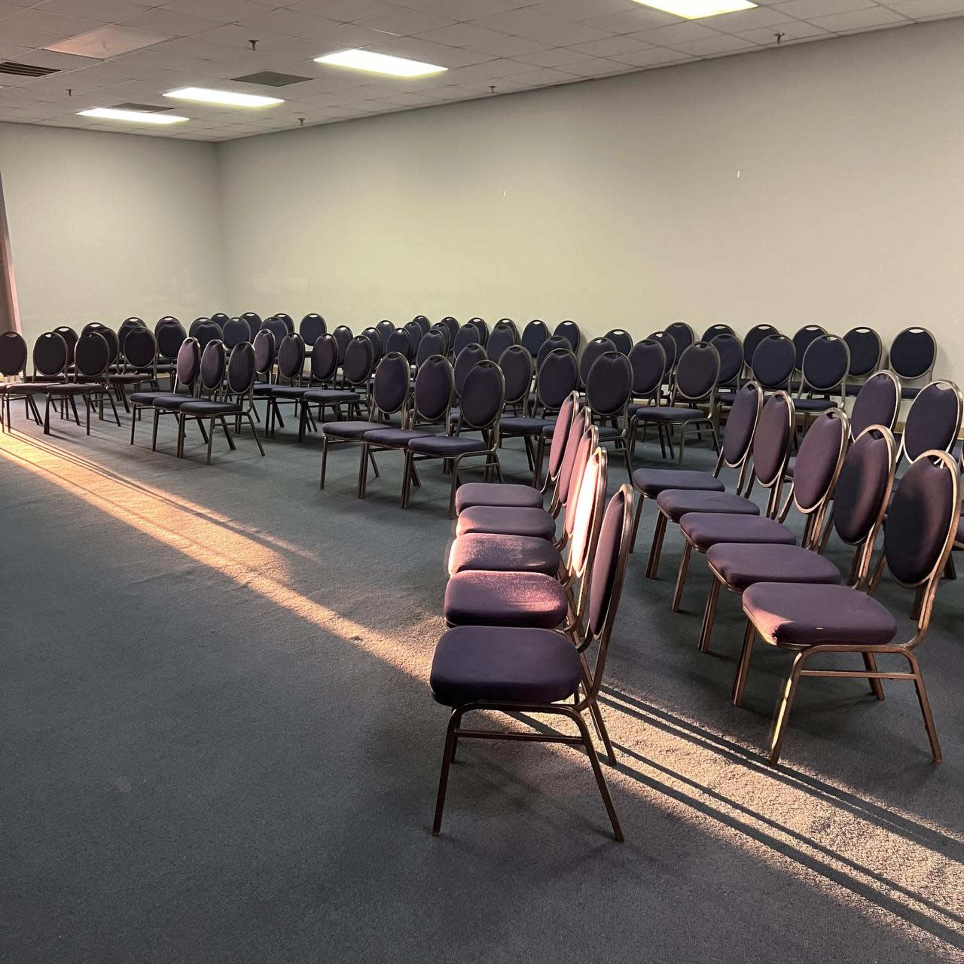 The image shows a large room with rows of empty, circular-backed chairs arranged neatly on a carpeted floor.