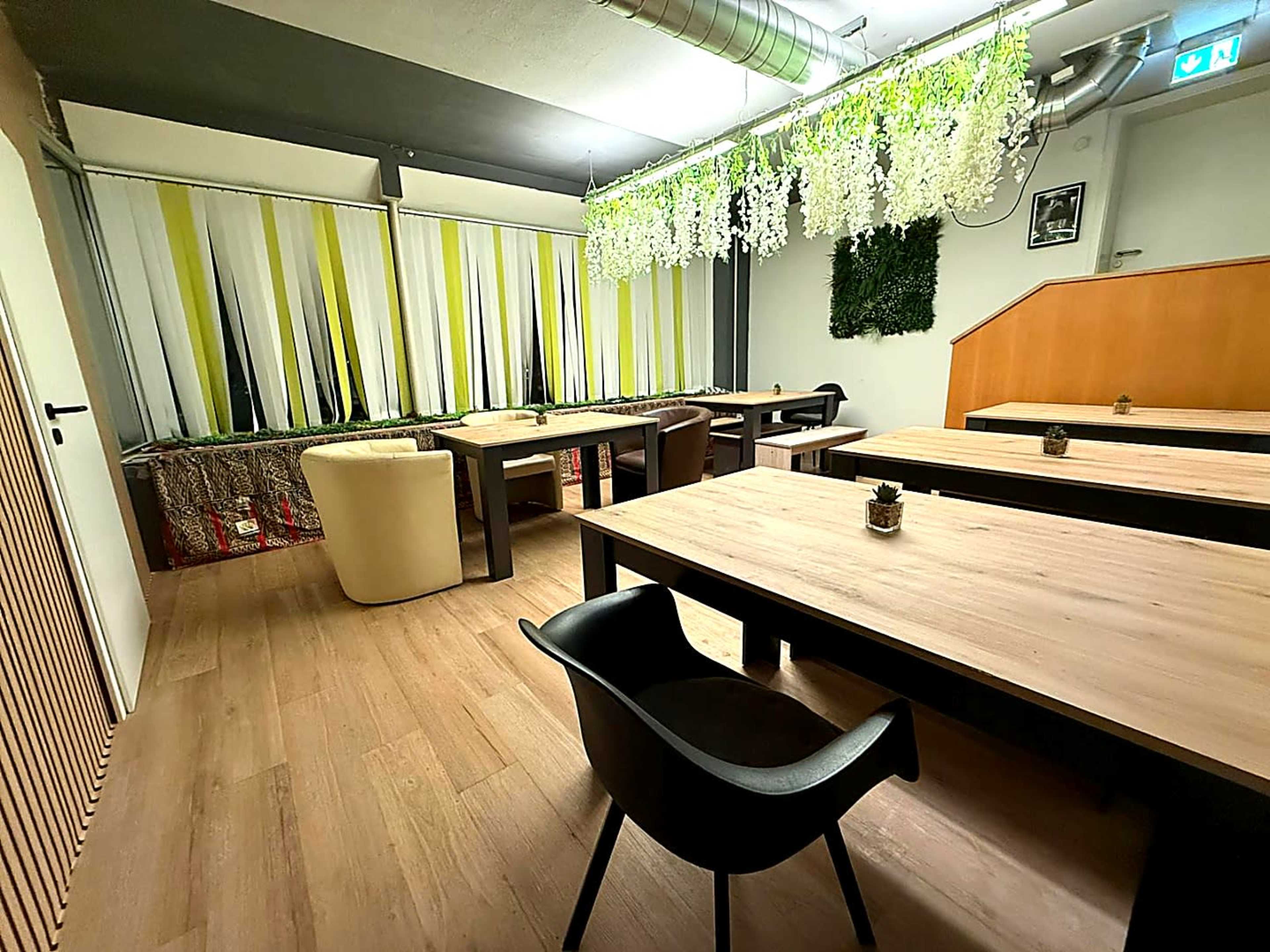 The image shows a modern dining area with wooden tables, minimalist chairs, and decorative plants hanging from the ceiling.