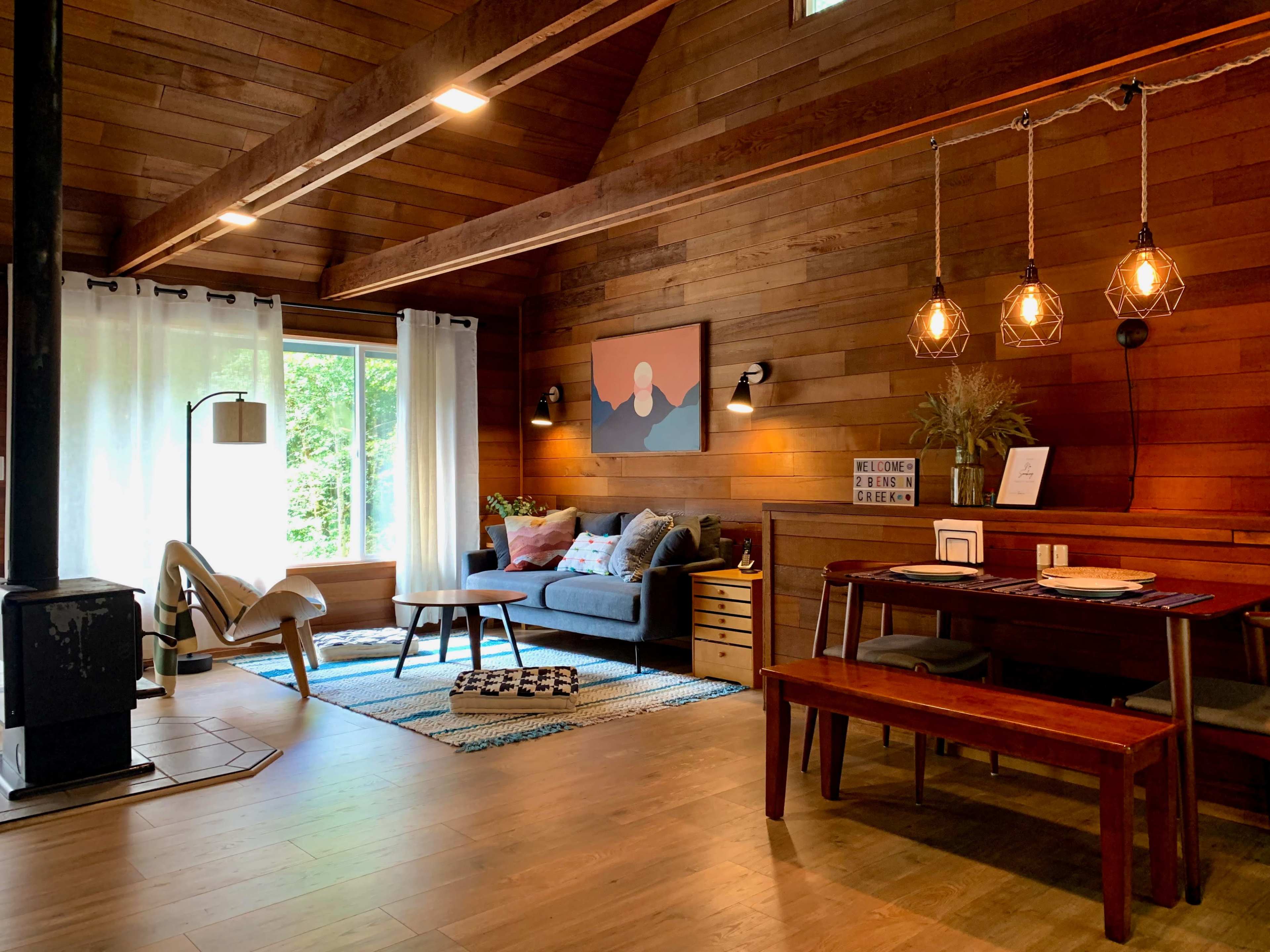 A cozy living room with wooden walls, large windows, a dining table, and a mix of seating areas.