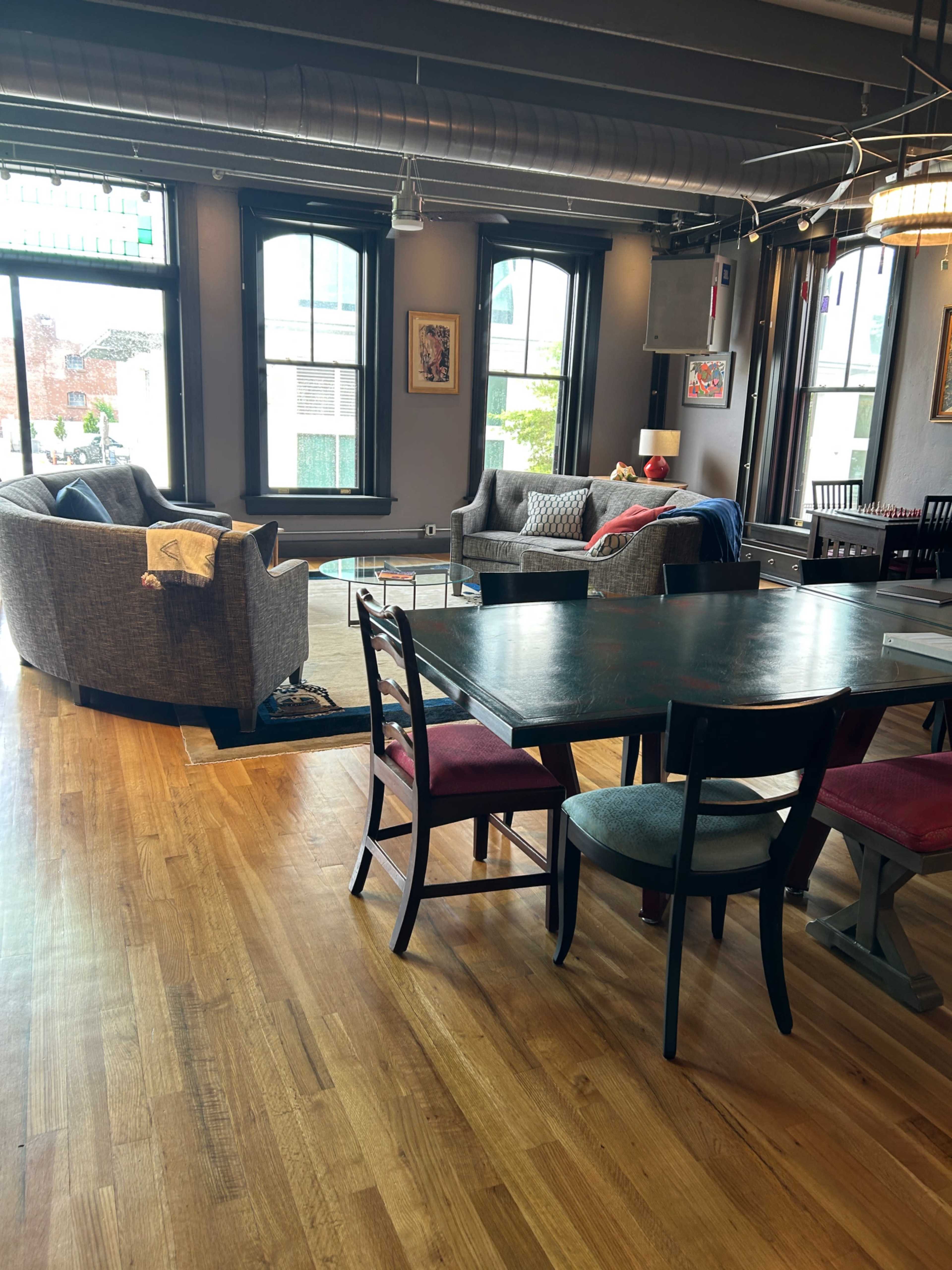 The image shows a modern living space with a dining table and chairs, a gray couch, and large windows allowing natural light to enter.