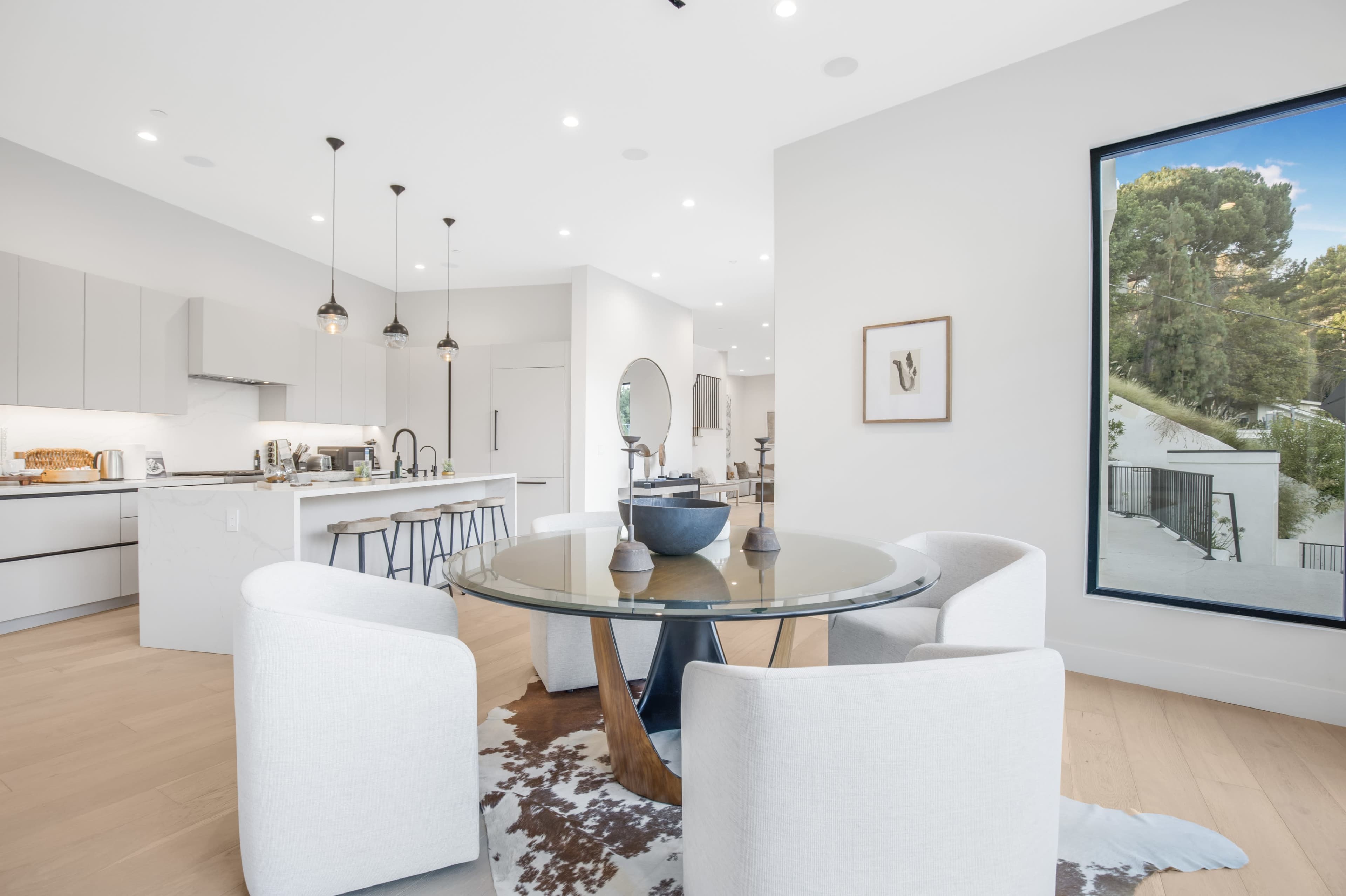 The image shows a modern dining area with a round glass table surrounded by white chairs, complemented by a spacious kitchen in the background.
