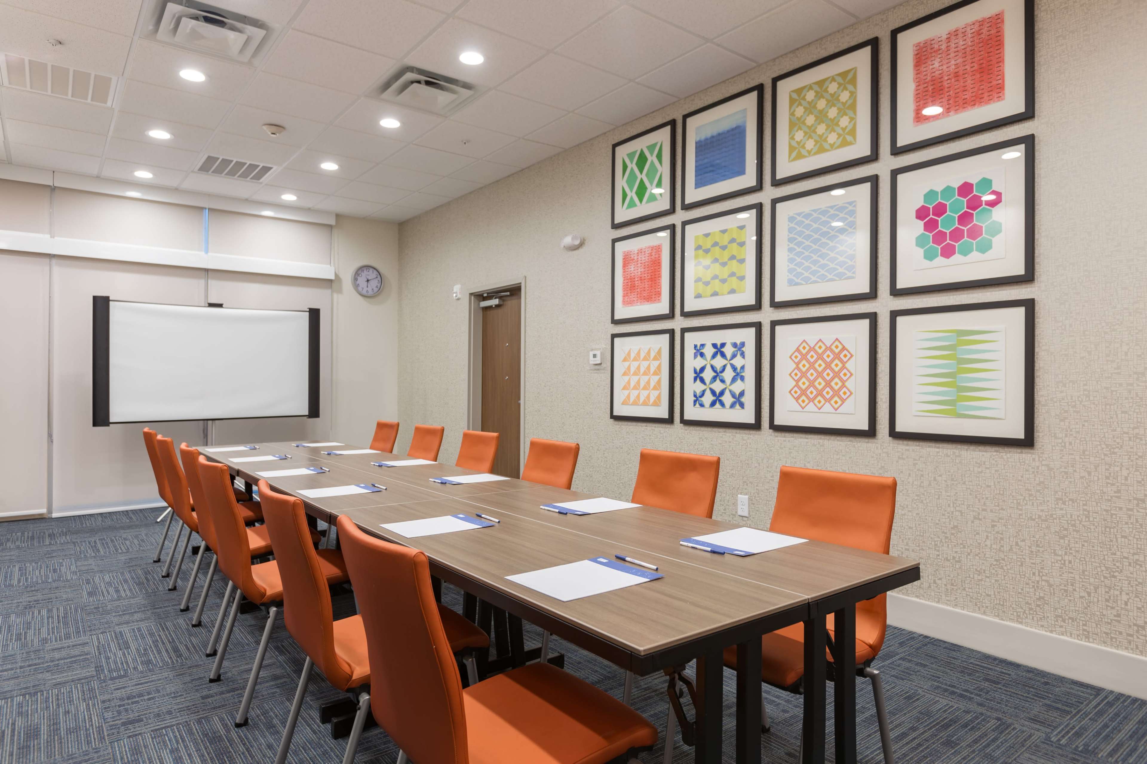 A conference room features a long wooden table with orange chairs, surrounded by colorful framed artwork on the walls and a projector screen at the front.