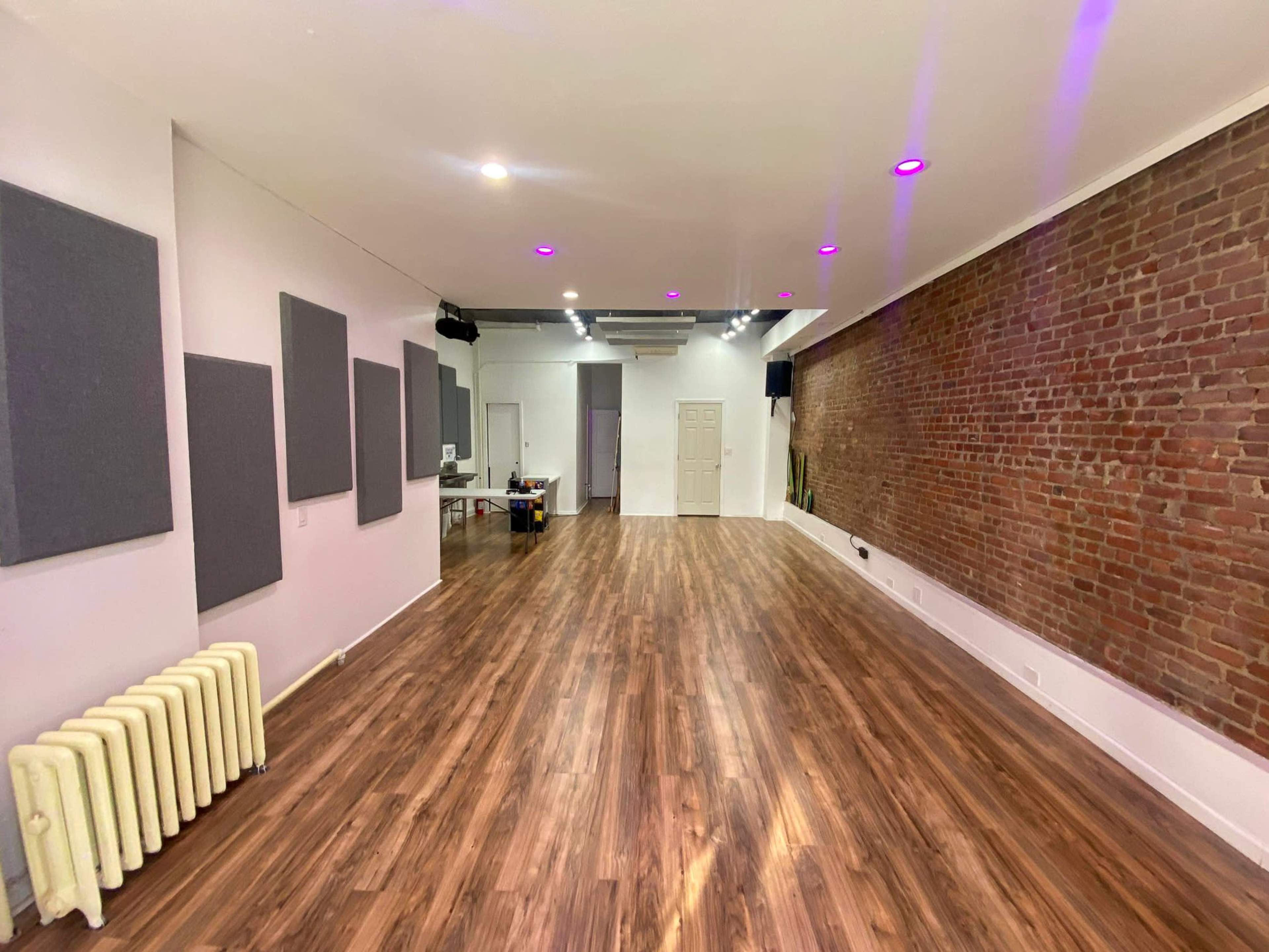 A spacious room with hardwood flooring, exposed brick walls, soundproof panels, and bright overhead lights.