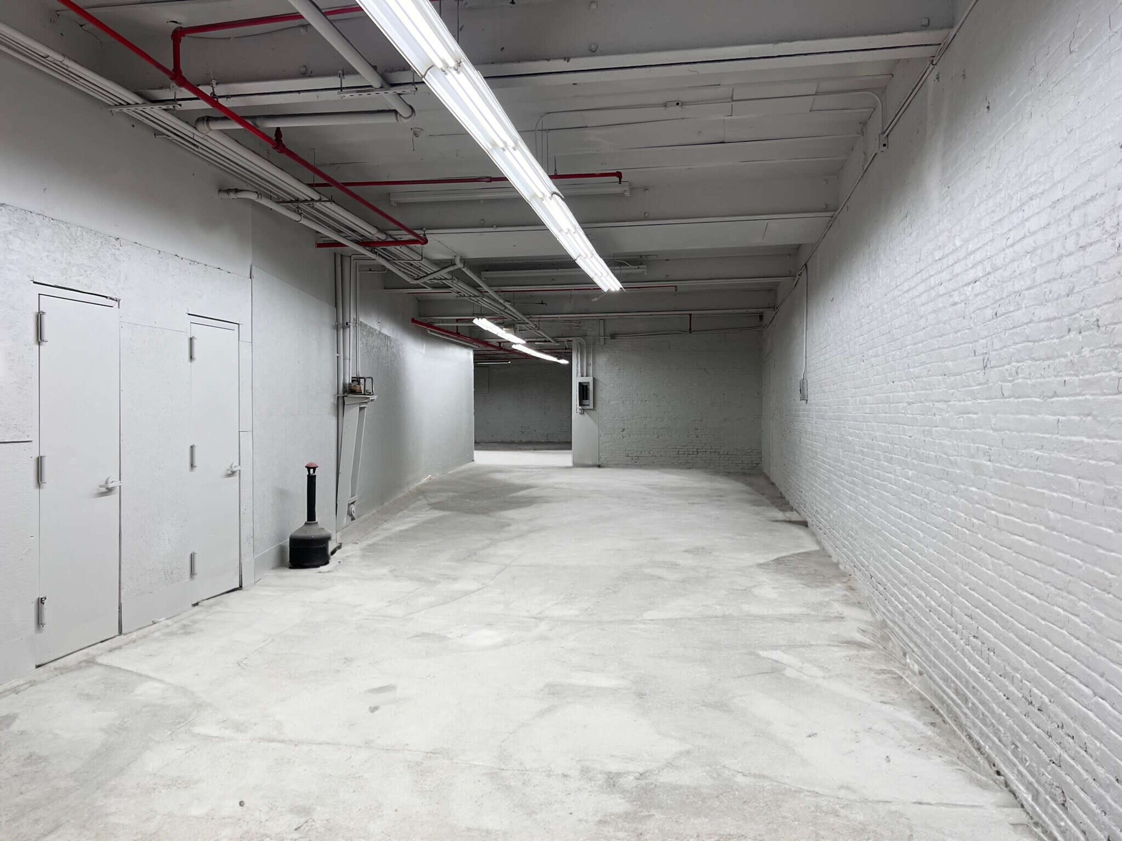The image shows a long, empty corridor with gray walls, concrete flooring, and overhead fluorescent lighting.