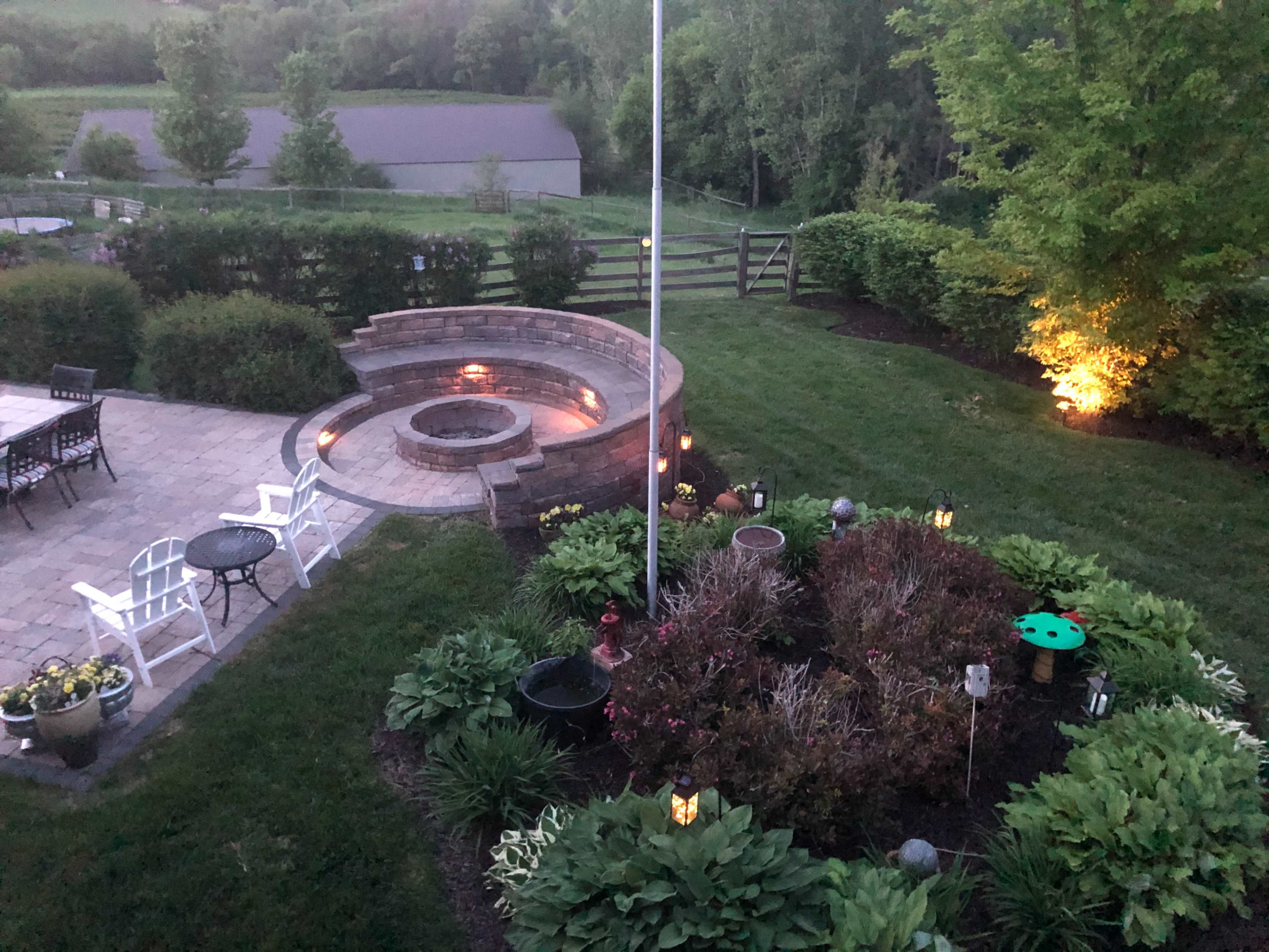 The image shows a landscaped garden with a circular stone fire pit, surrounded by flowers and greenery, and outdoor seating areas, illuminated by lights at dusk.