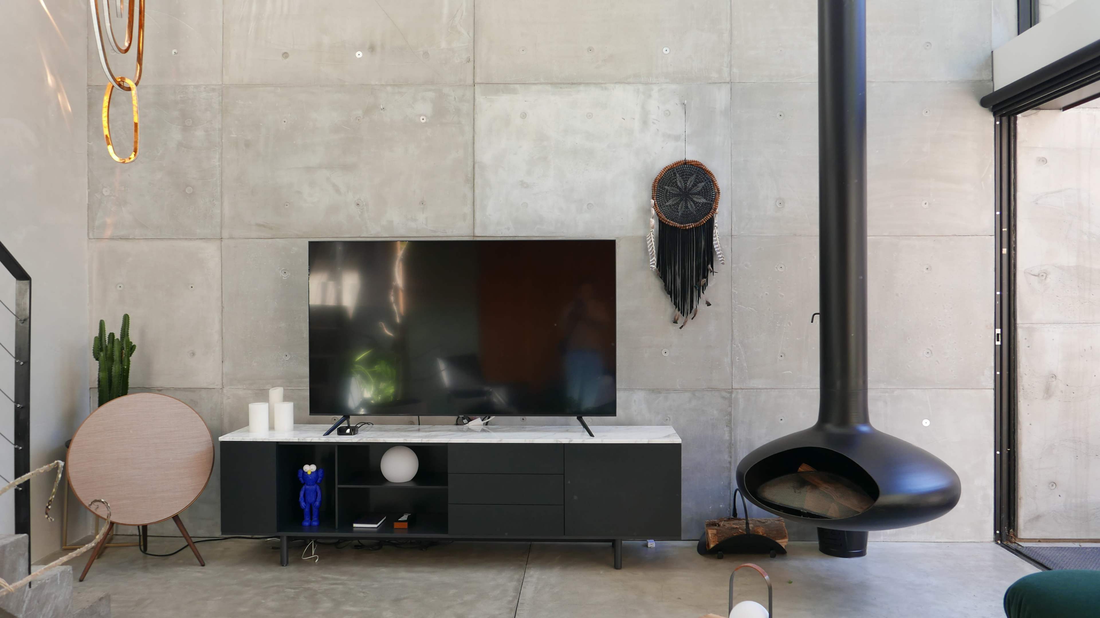 The image shows a living room with a concrete wall, a large flat-screen TV on a black media console, and a modern fireplace hanging from the ceiling.