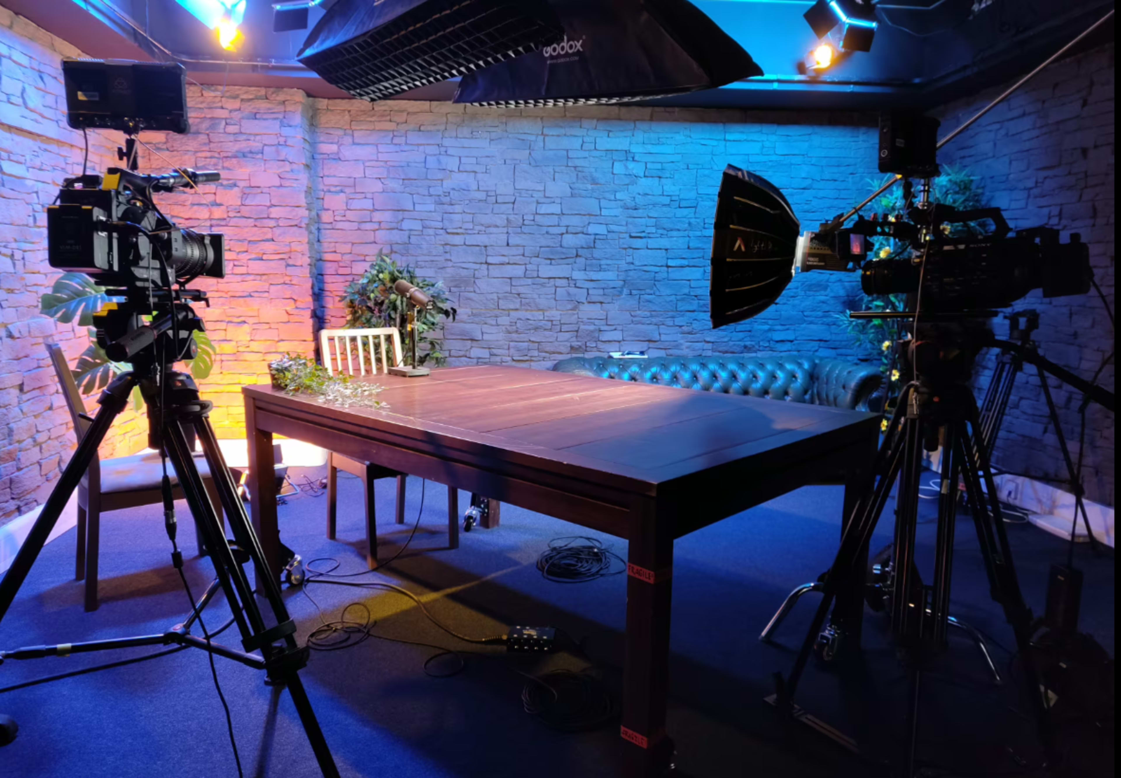 The image shows a dimly lit studio with a wooden table at the center, surrounded by three cameras and decorative plants against a stone wall backdrop.