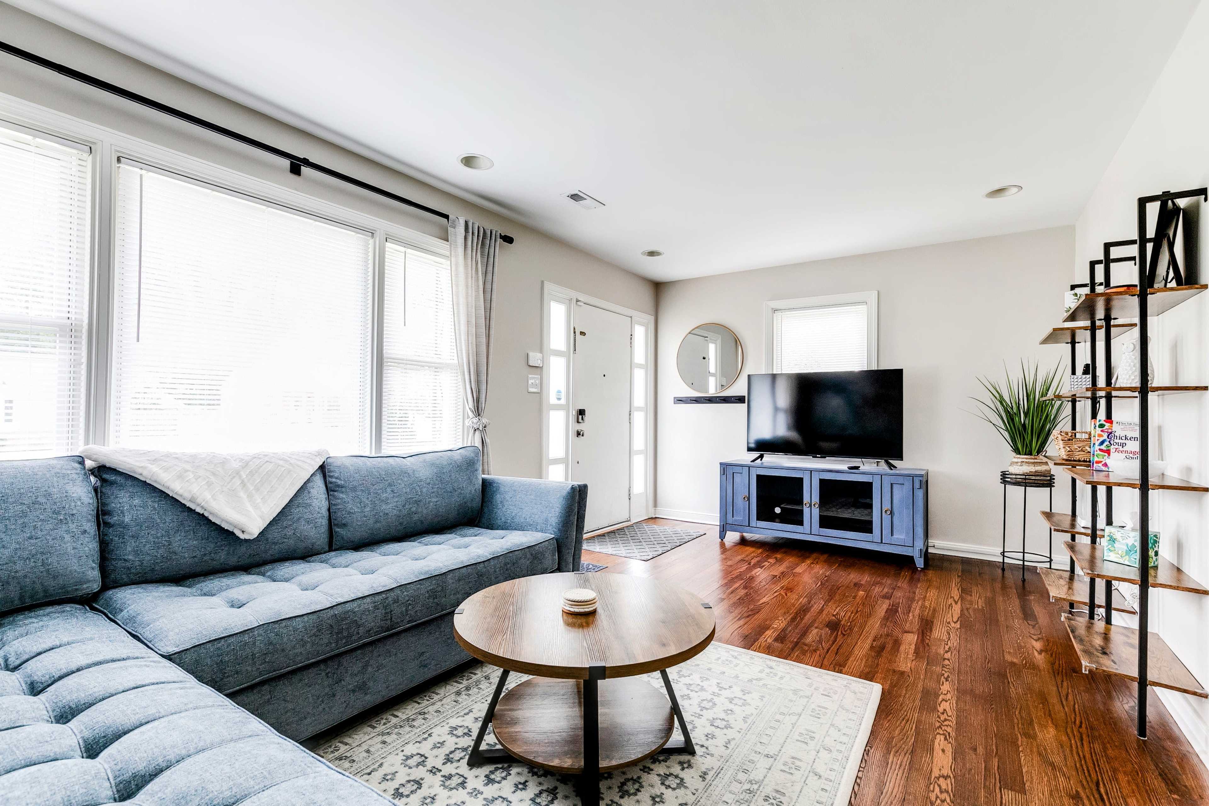 A modern living room features a blue sectional sofa, a round wooden coffee table, a television on a stand, and a mix of natural light from large windows.