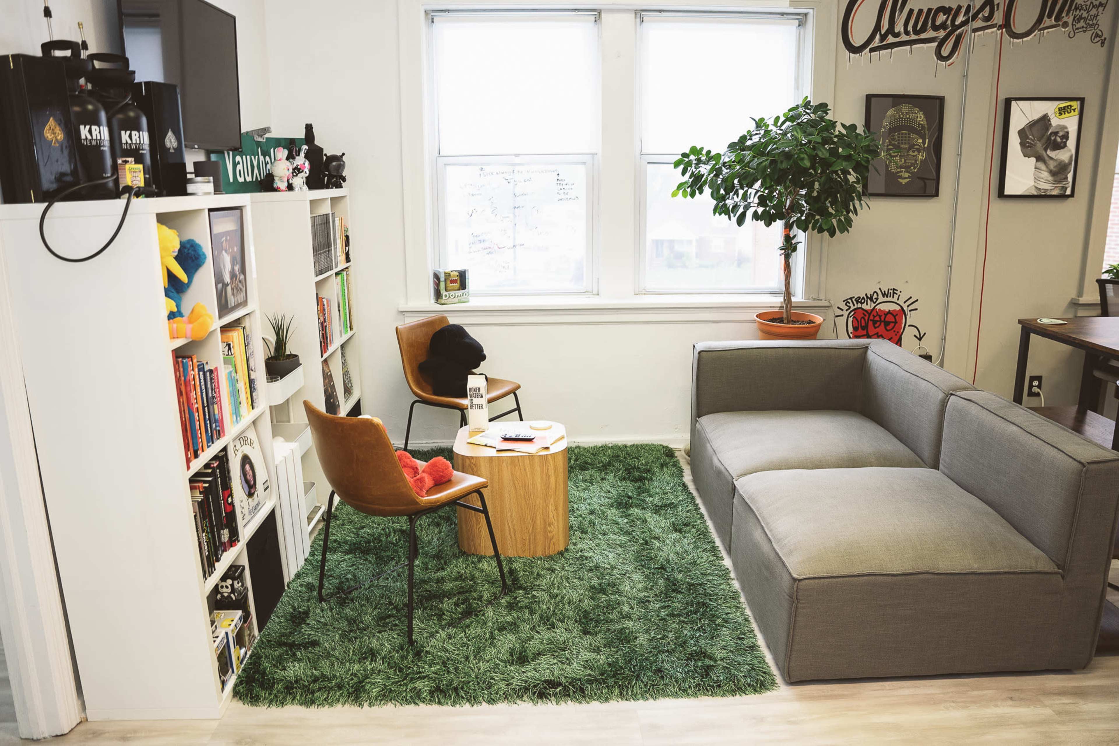 A cozy corner of a room with a grey couch, two chairs, a wooden coffee table, bookshelves filled with books, and a potted plant in a bright, green carpeted area.