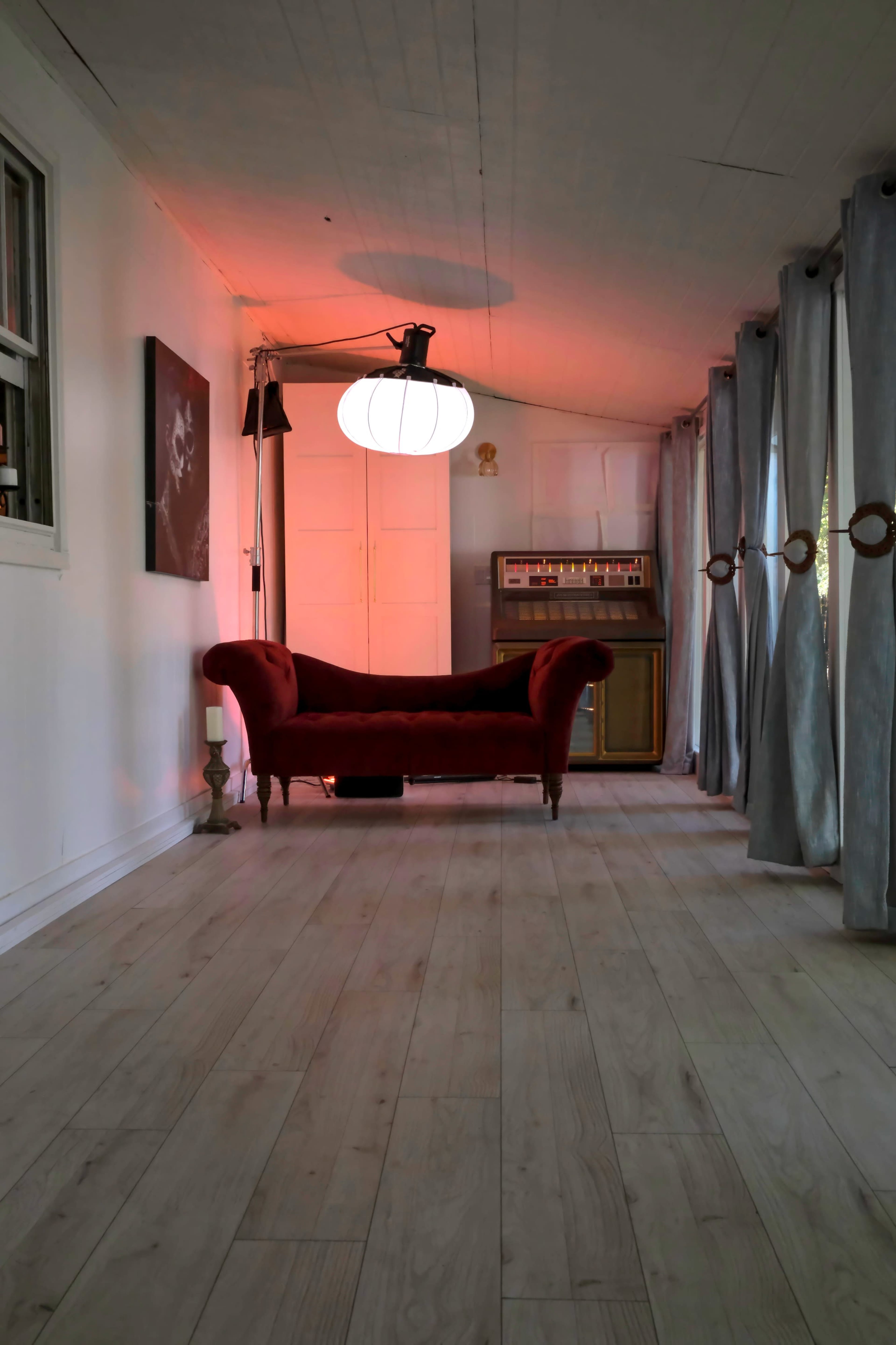 A red velvet couch is positioned in a well-lit room with wooden flooring, featuring a vintage record player and sheer curtains.