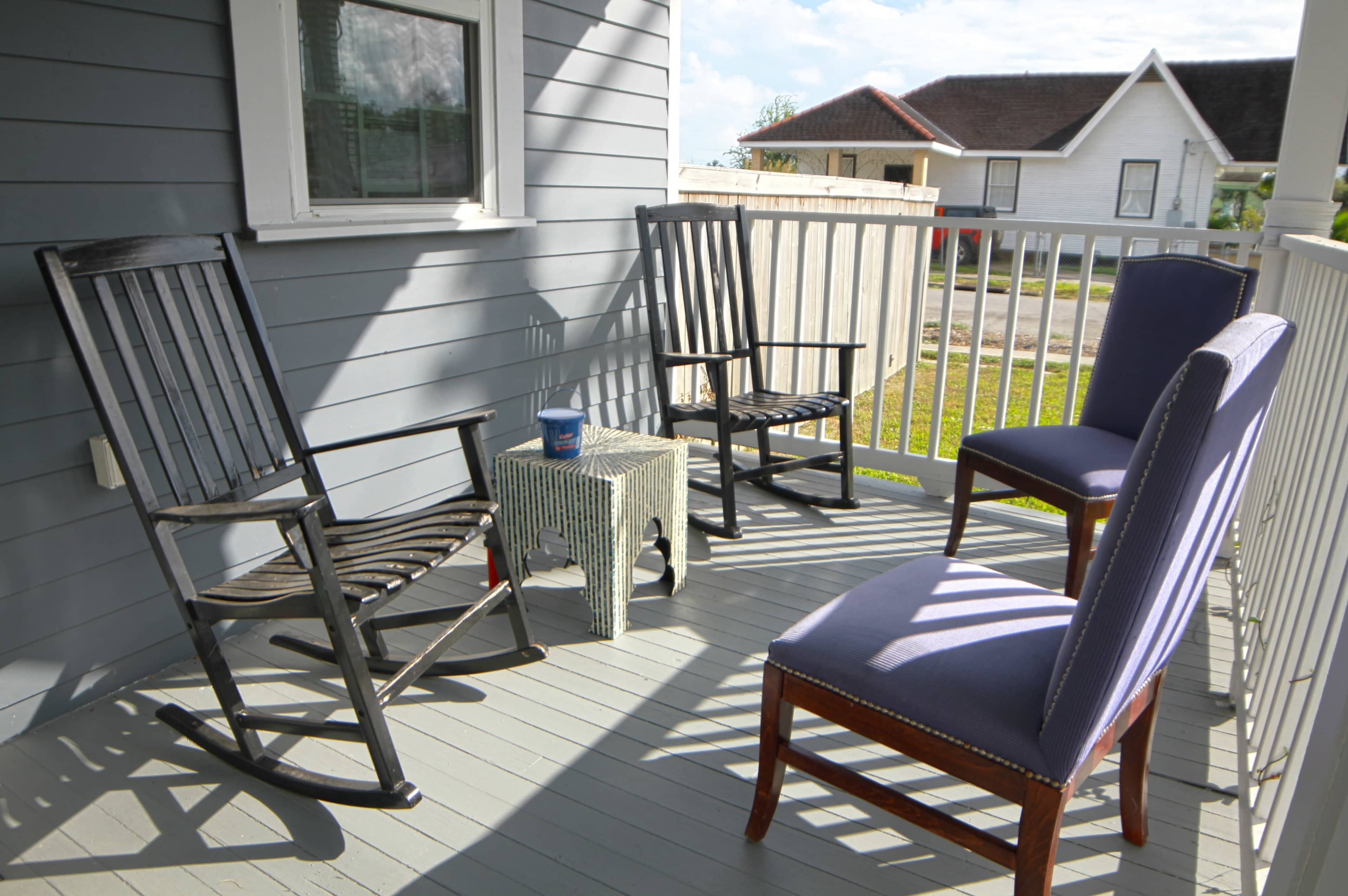 The image shows a porch with two rocking chairs, a small table, and two upright chairs against a backdrop of a nearby house and green lawn.