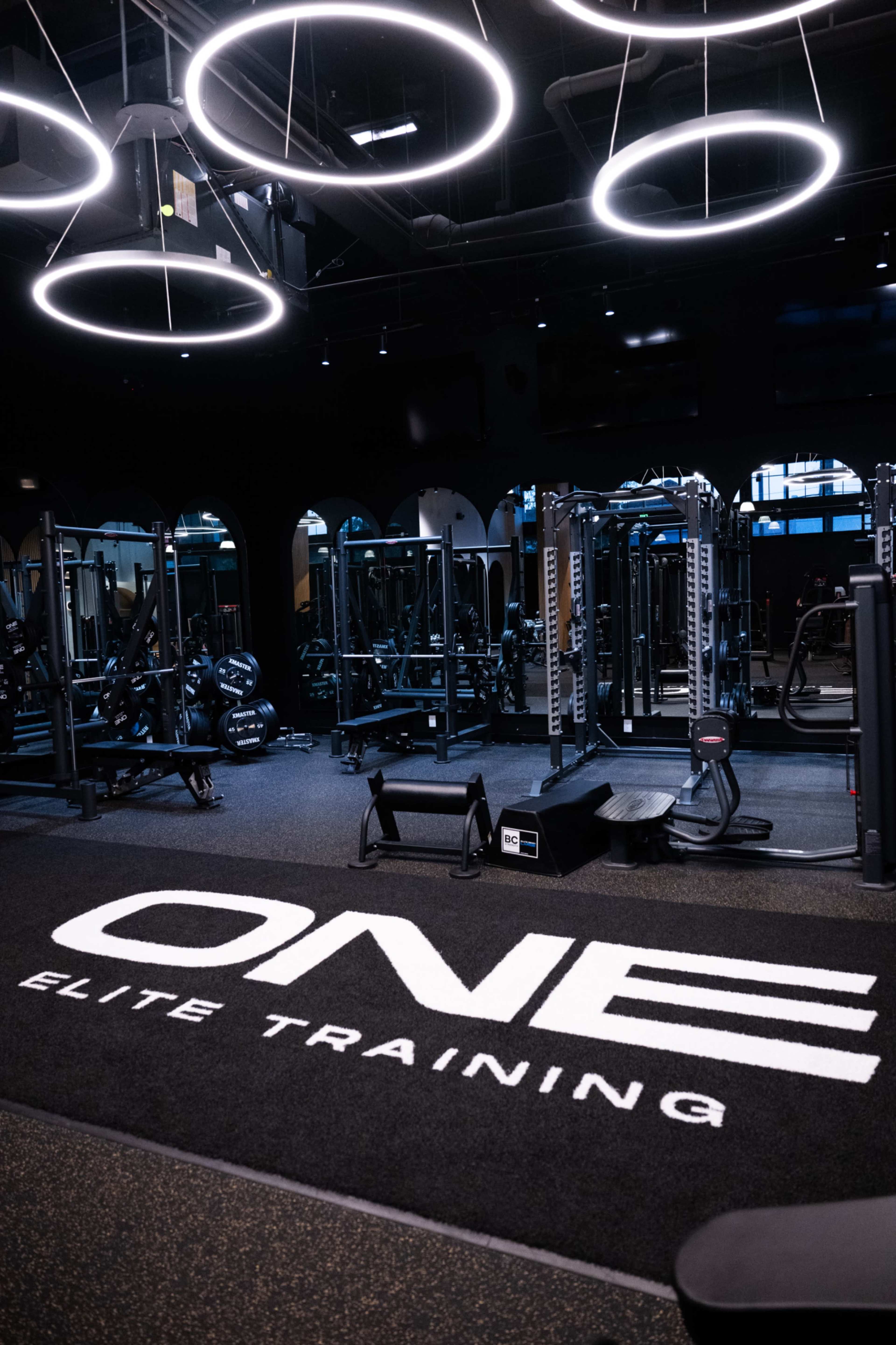 The image shows a modern gym interior with a black carpet featuring the words "ONE ELITE TRAINING," illuminated by circular overhead lights and equipped with various workout machines.