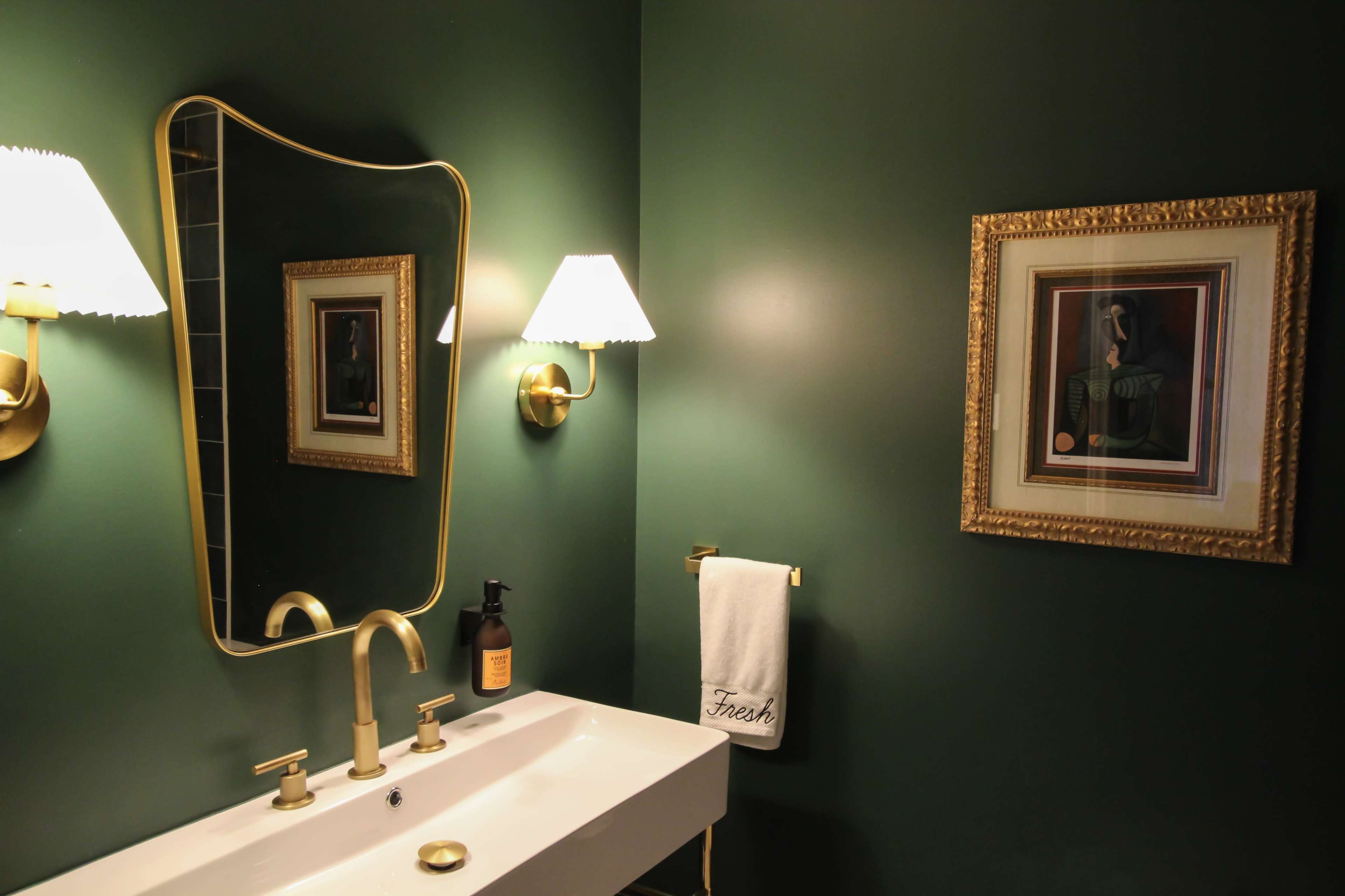 The image shows a bathroom with dark green walls, a decorative mirror, a gold-framed artwork, and a white towel hung beside a modern sink with gold fixtures.