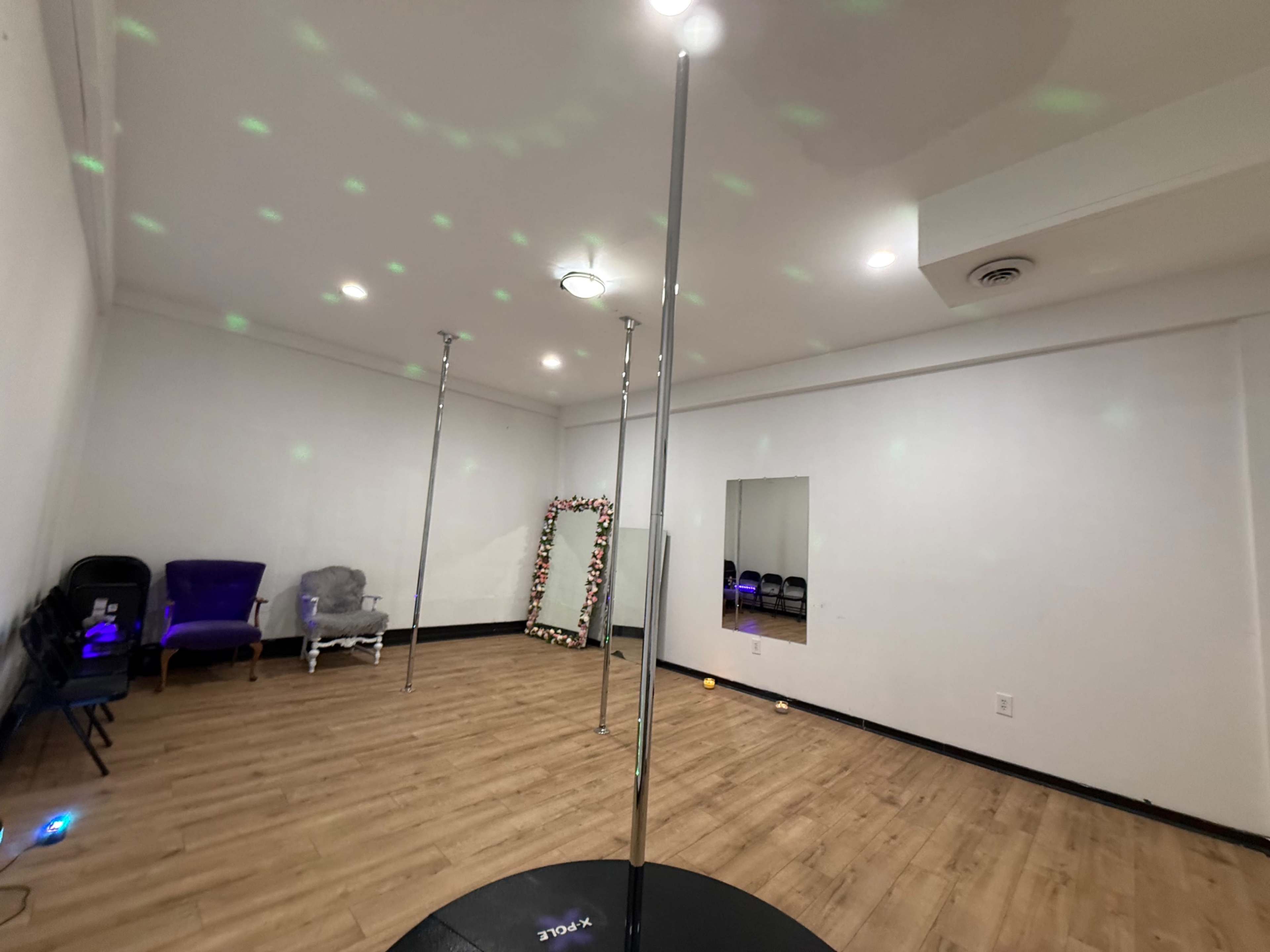 The image shows a spacious room with two metallic poles, a large mirror, and a few pieces of furniture including chairs and a couch.