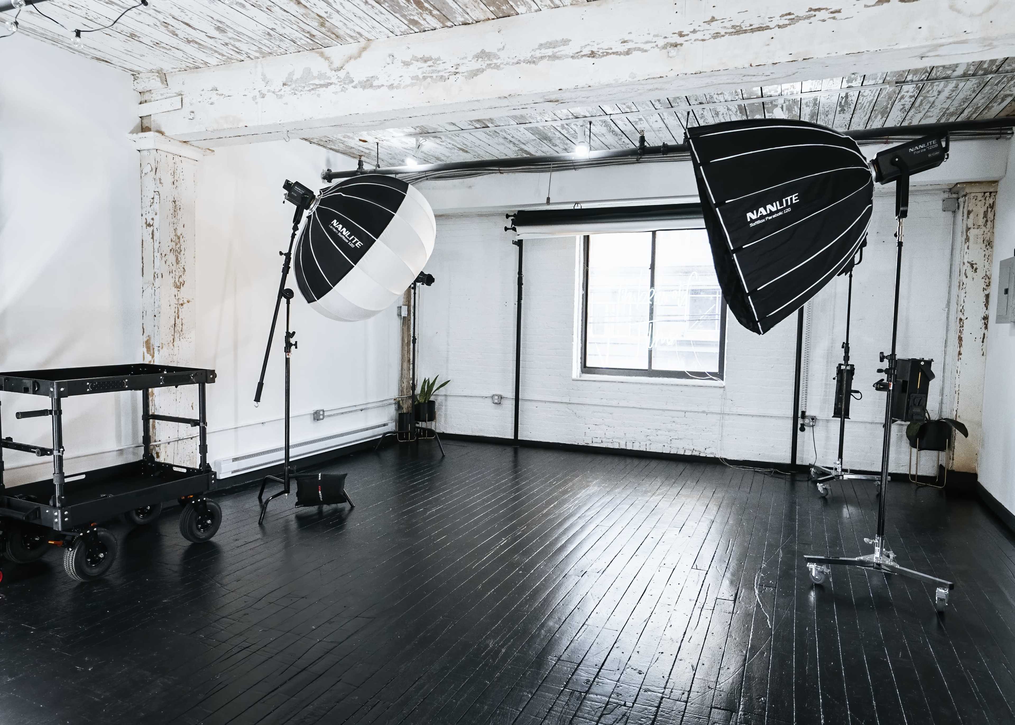 A photography studio with two softbox lights, a black cart, and a large window.