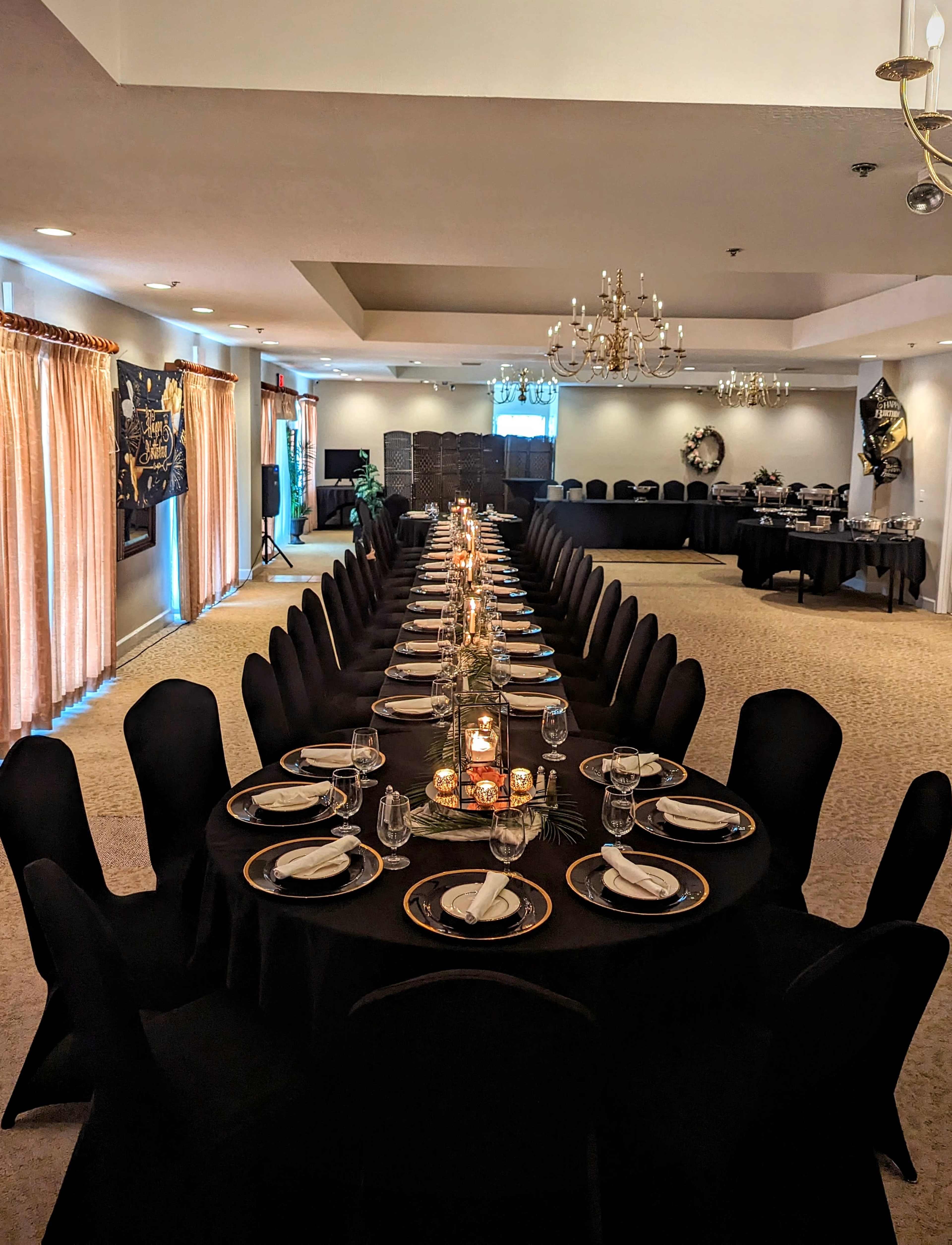 A long banquet table is elegantly set with black tablecloths, gold-rimmed plates, and candles, surrounded by decorated chairs in a well-lit event space.