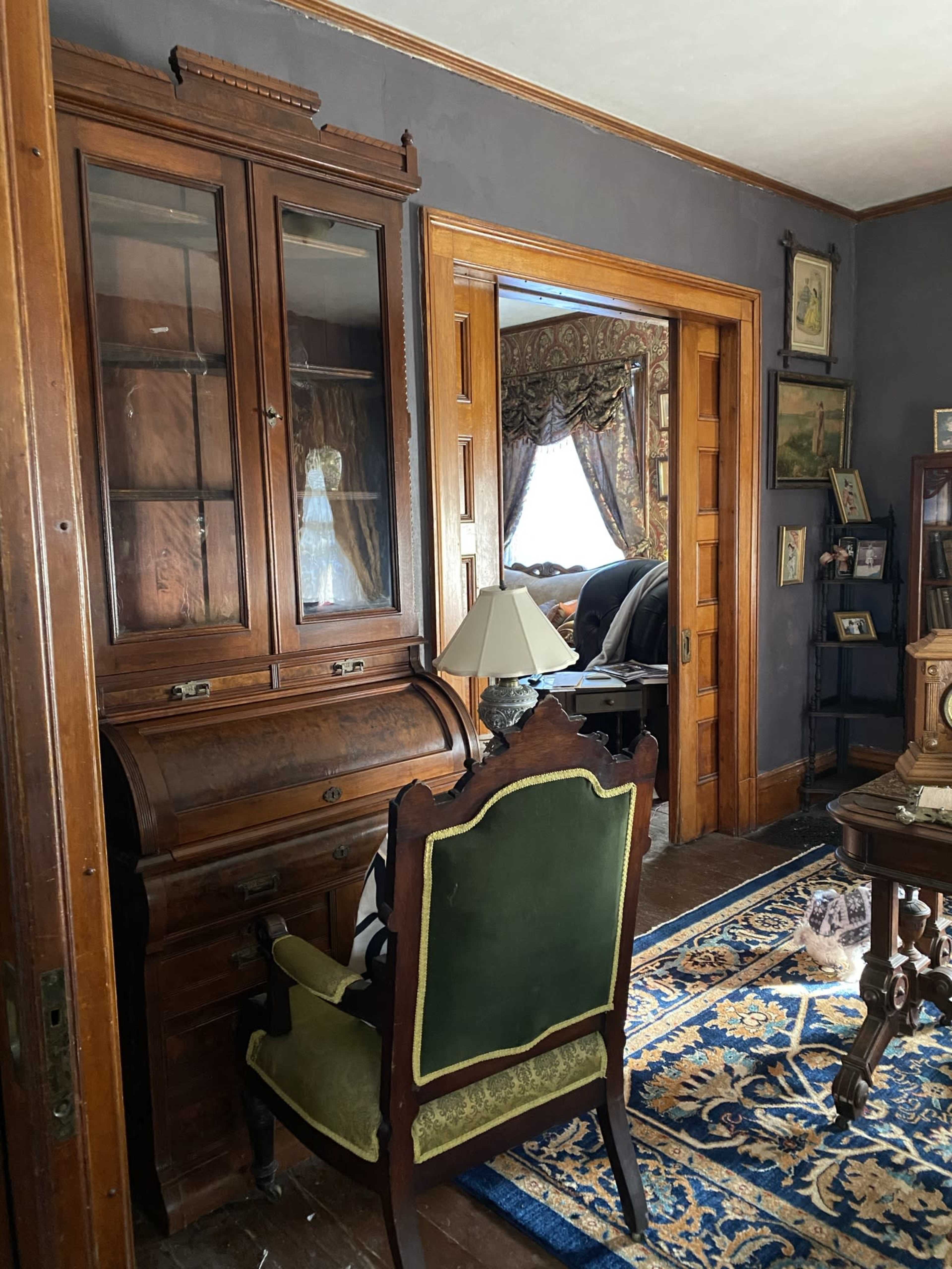 A room featuring a wooden desk with a green upholstered chair, a lamp, and framed pictures on the walls, with a doorway leading to another room adorned with curtains.