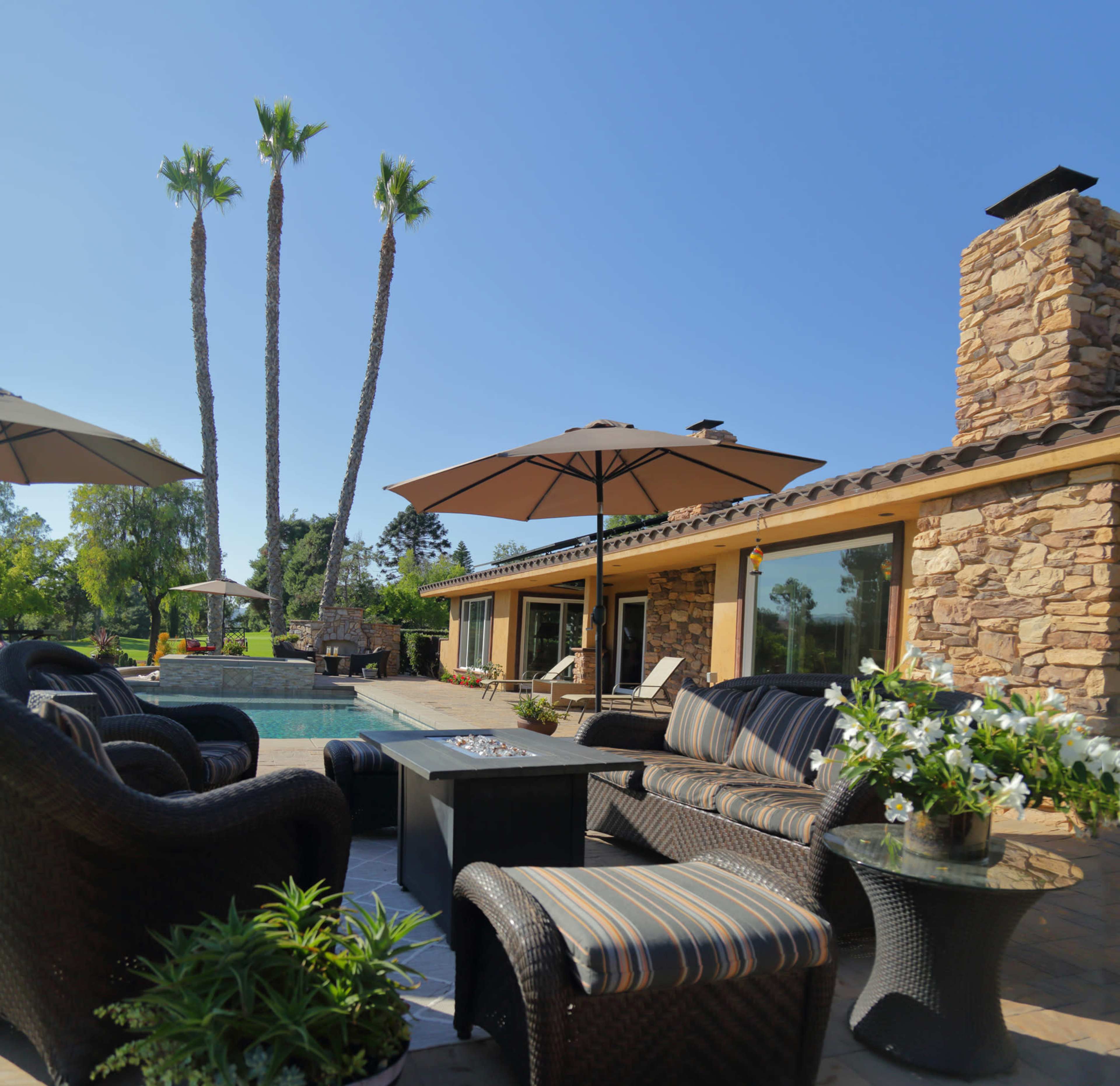 A patio area with seating and umbrellas surrounds a swimming pool, accompanied by palm trees and a stone house in the background.