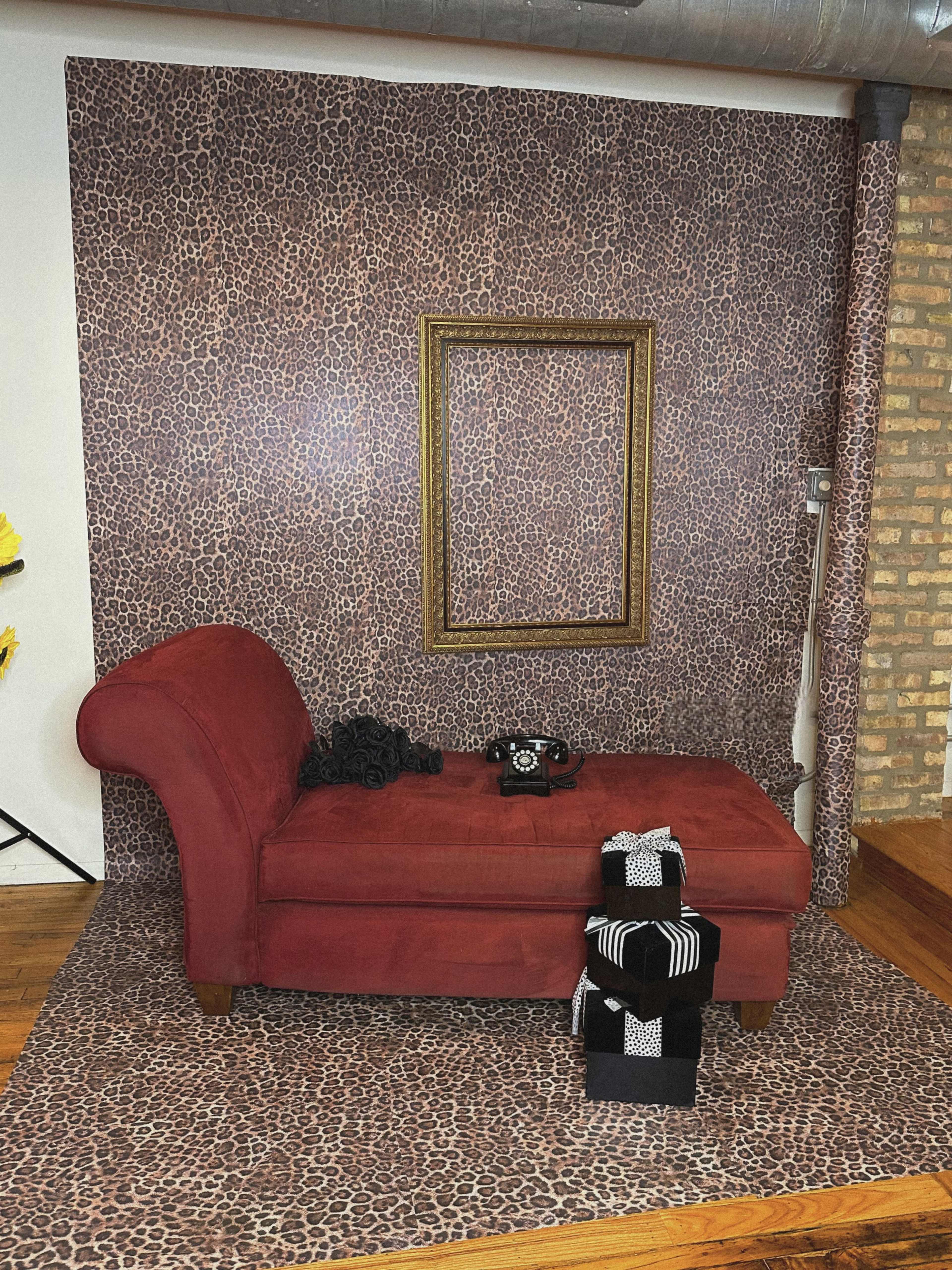 The image features a red chaise lounge positioned in front of a leopard print wall, accompanied by a gold frame and several black and white gift boxes on the floor.