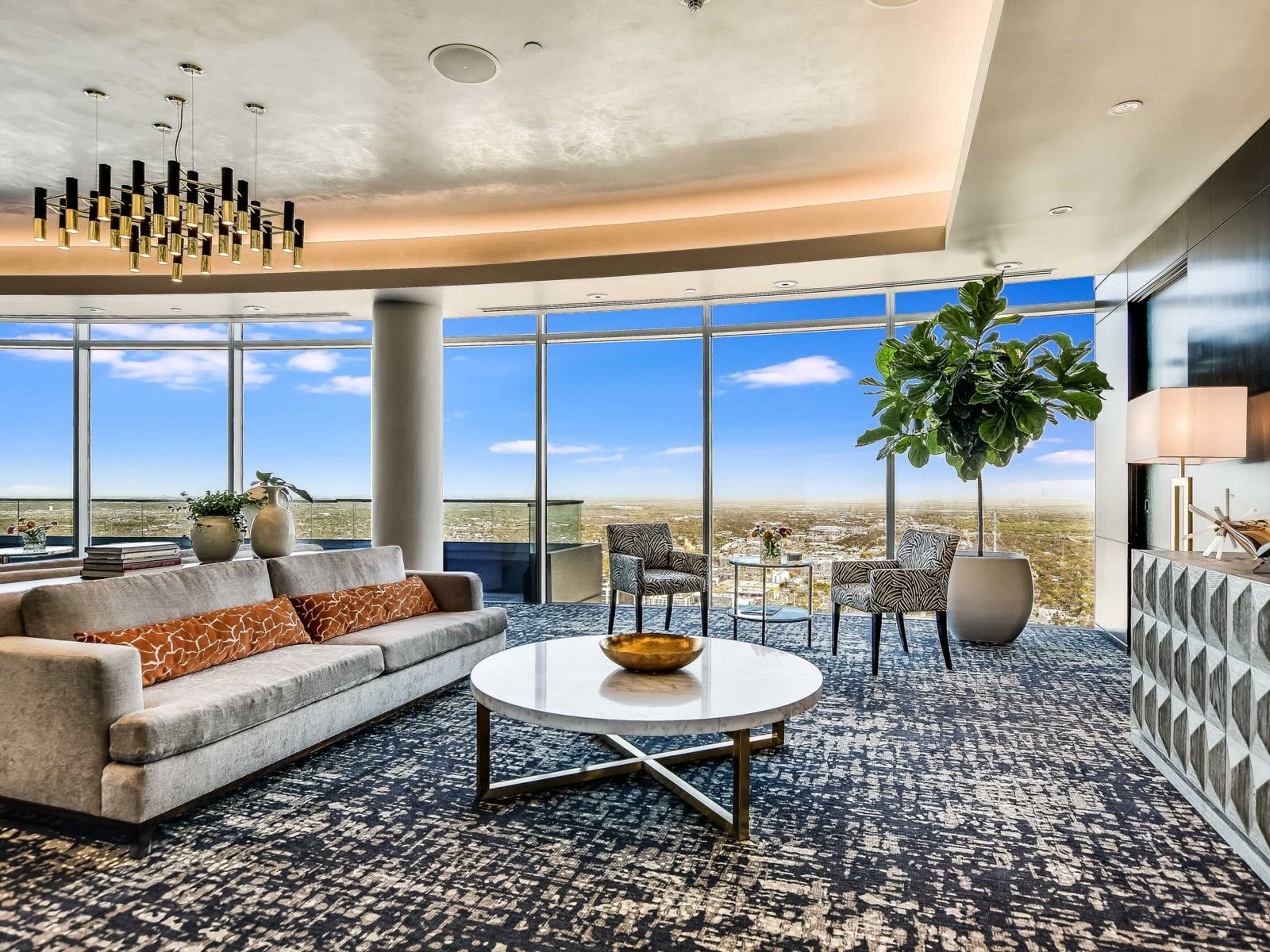 A modern living room features a gray sofa with decorative pillows, a round coffee table, two chairs, and large windows showcasing a panoramic view of the landscape.