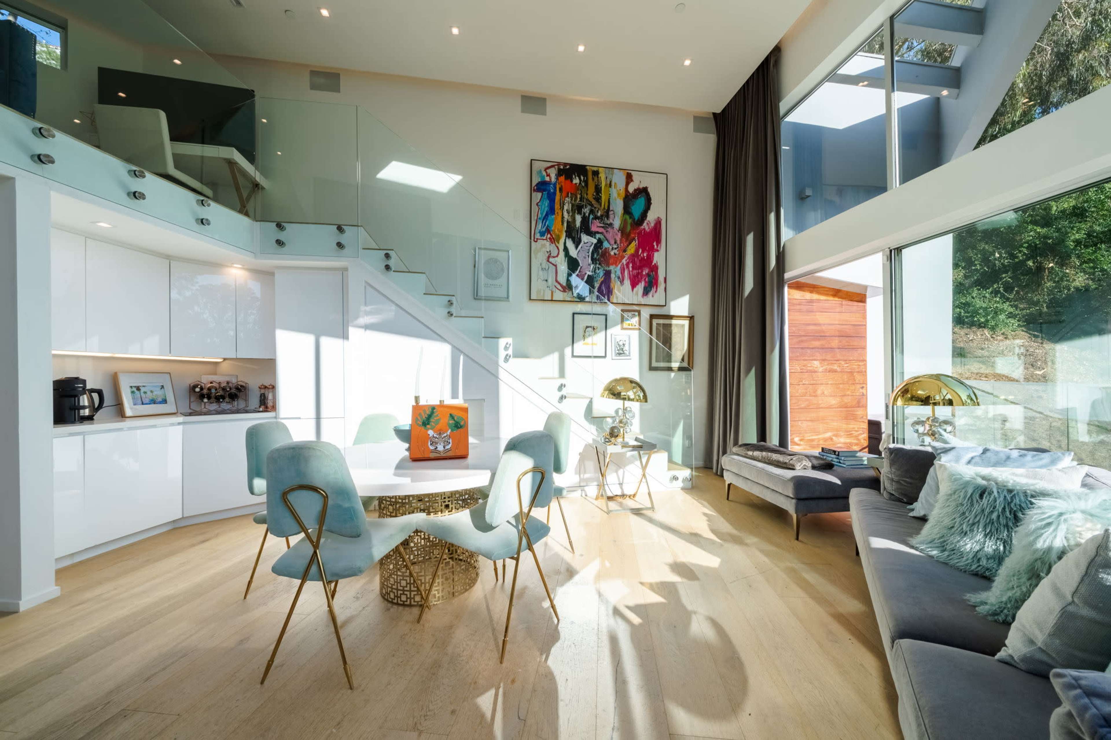 A modern interior featuring a dining area with a round table and chairs, a staircase with glass railings, and large windows allowing natural light to fill the space.
