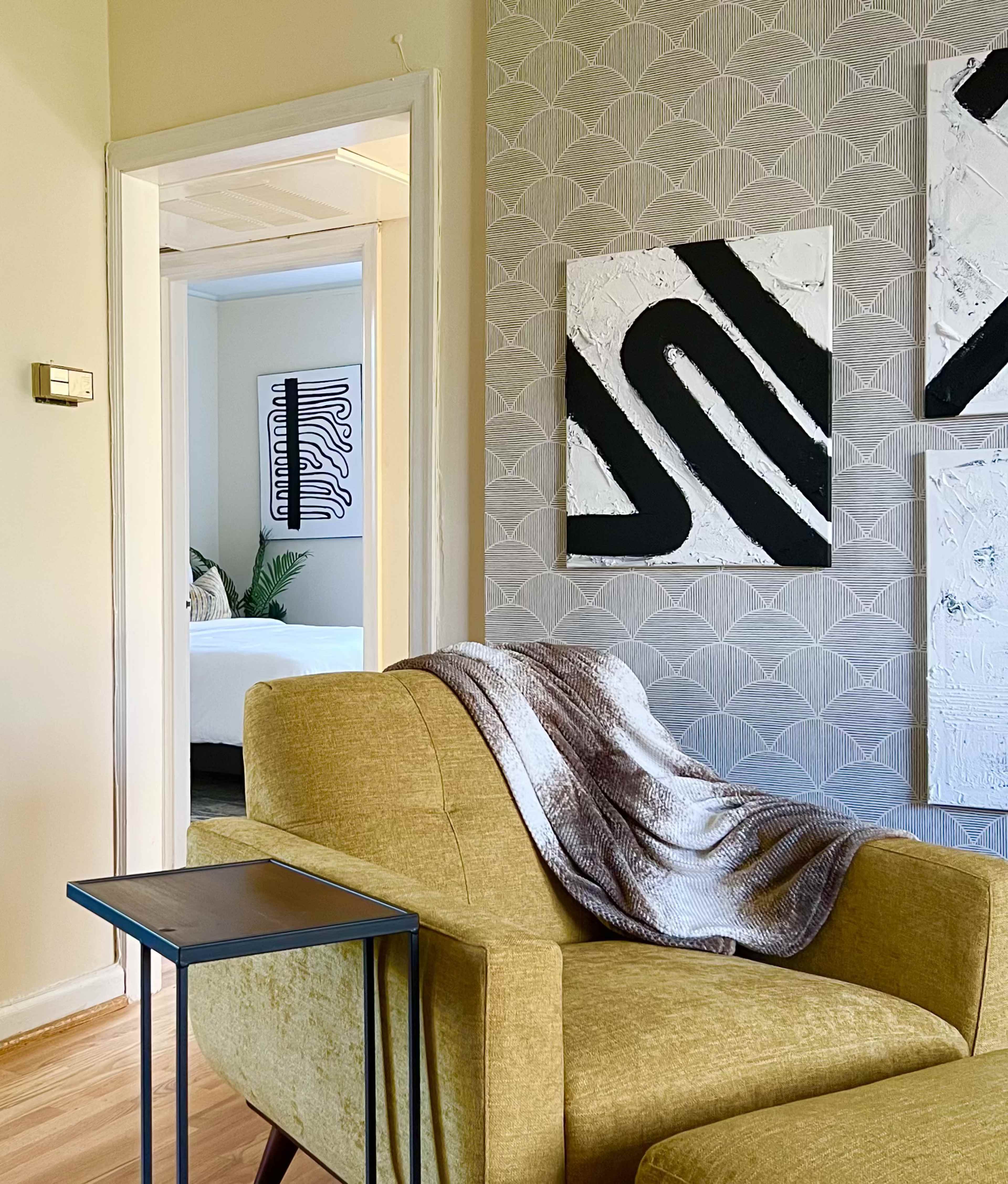 A yellow armchair with a patterned throw blanket sits beside a small table in a room featuring abstract wall art and a glimpse of a bedroom through an open doorway.
