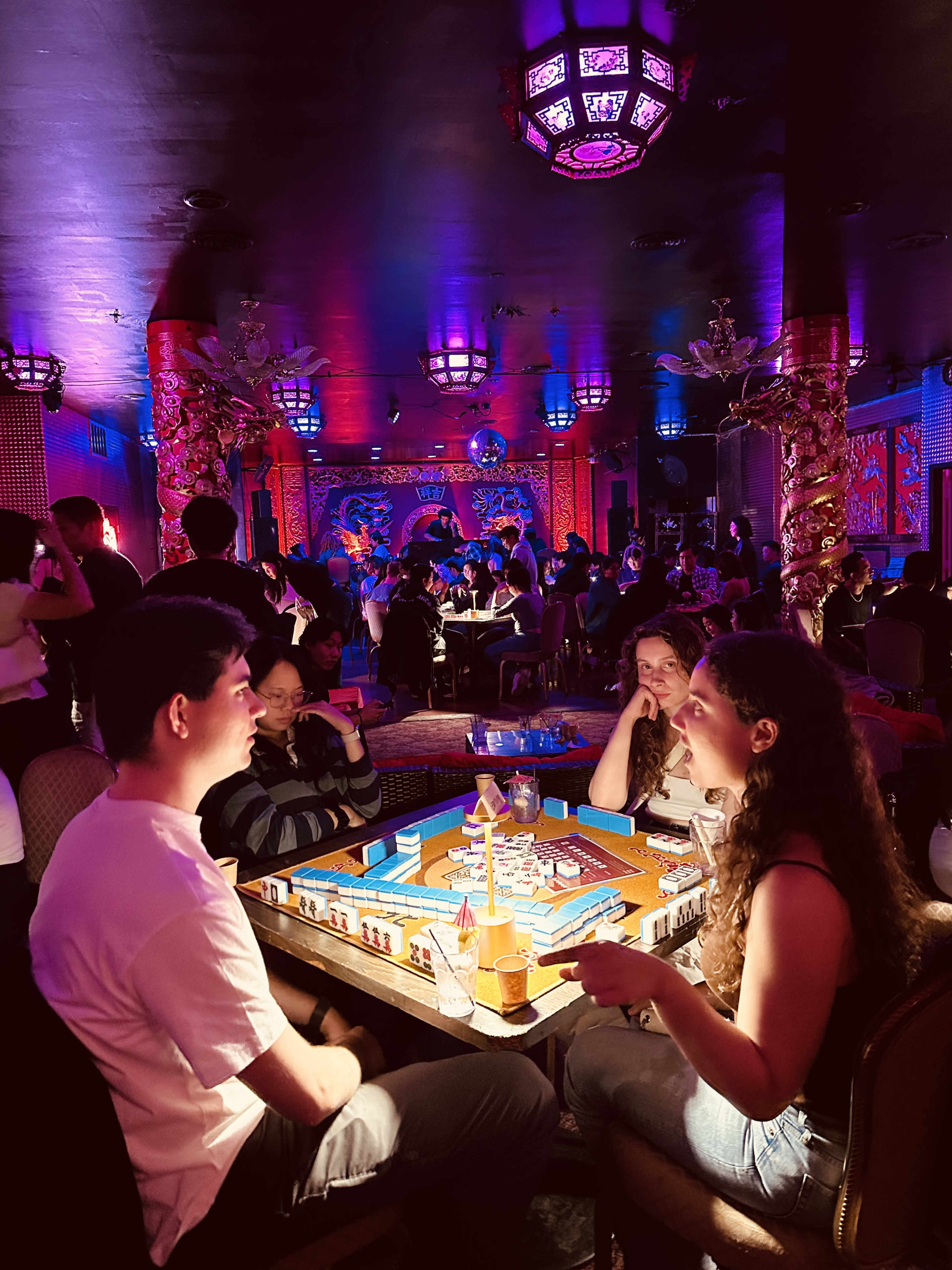 A lively indoor scene in a vibrant lounge features two people engaged in a board game, surrounded by groups of patrons enjoying their evening.
