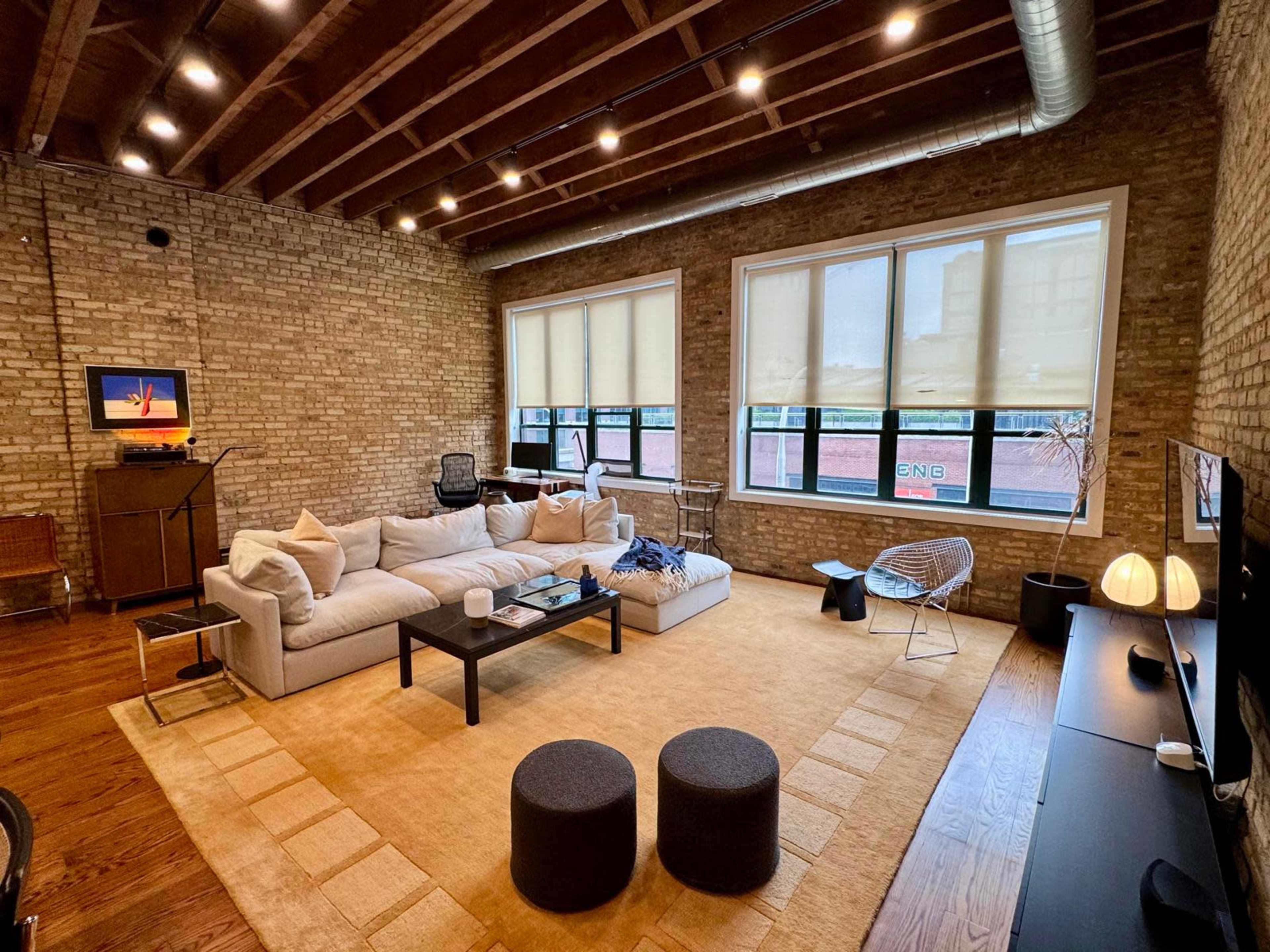 The image shows a spacious living room with exposed brick walls, large windows, a sectional sofa, a coffee table, and a television setup on a wooden floor.