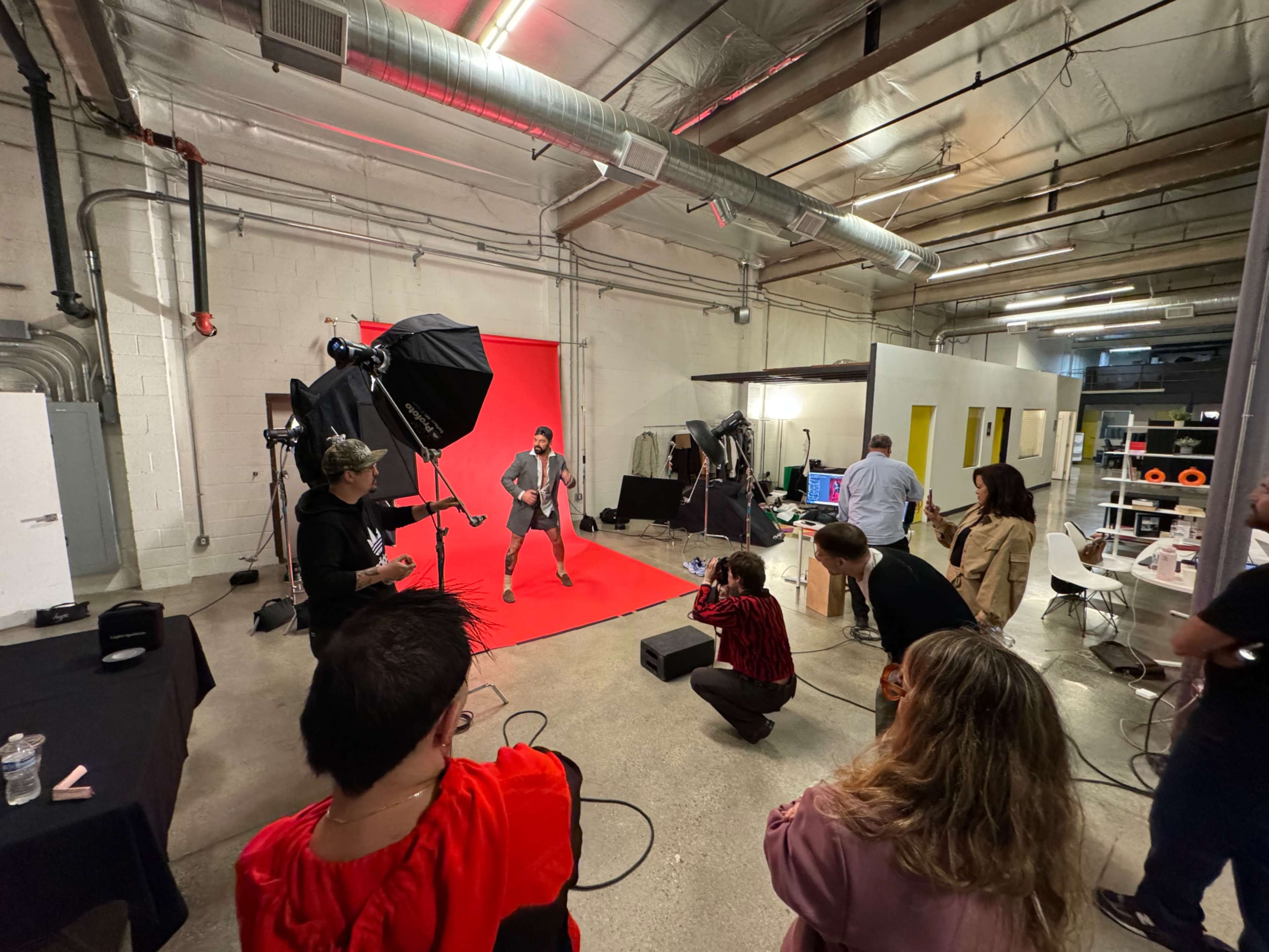 A group of people is gathered in a studio with a red backdrop, where one person is posing for a photo shoot while others observe and assist.