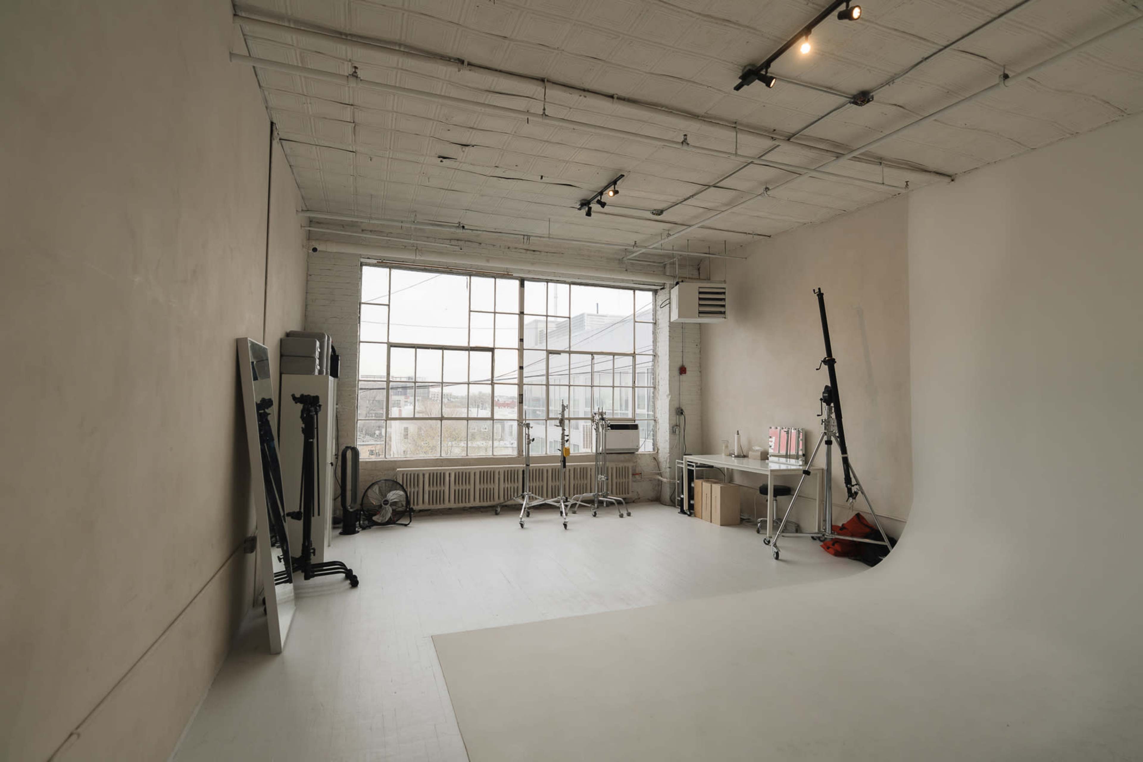 The image shows a spacious, well-lit photography studio with large windows, a seamless backdrop, and various equipment along the walls.
