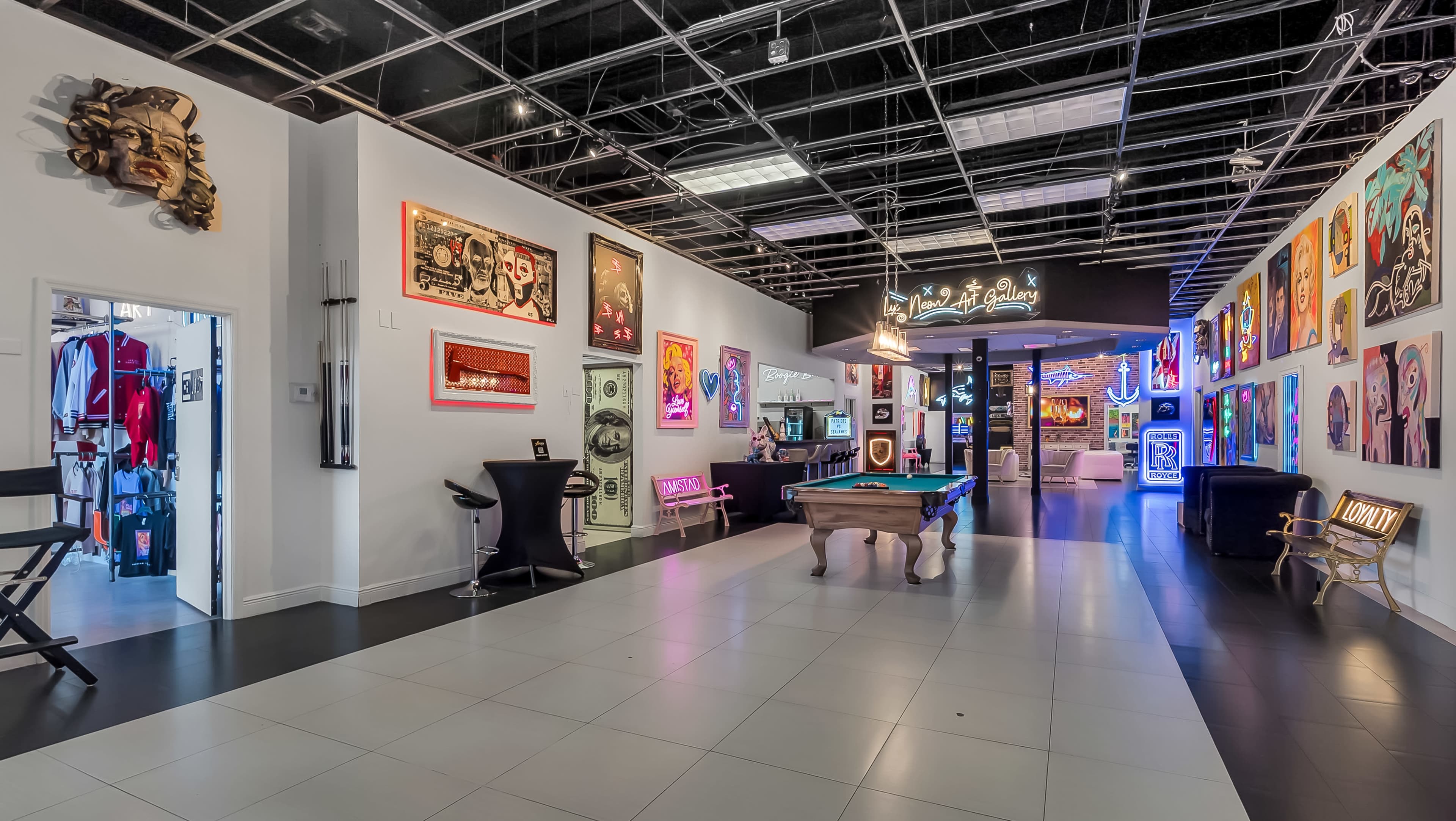 The image features a spacious art gallery with vibrant wall art, a pool table in the center, and an illuminated entrance leading to another area.
