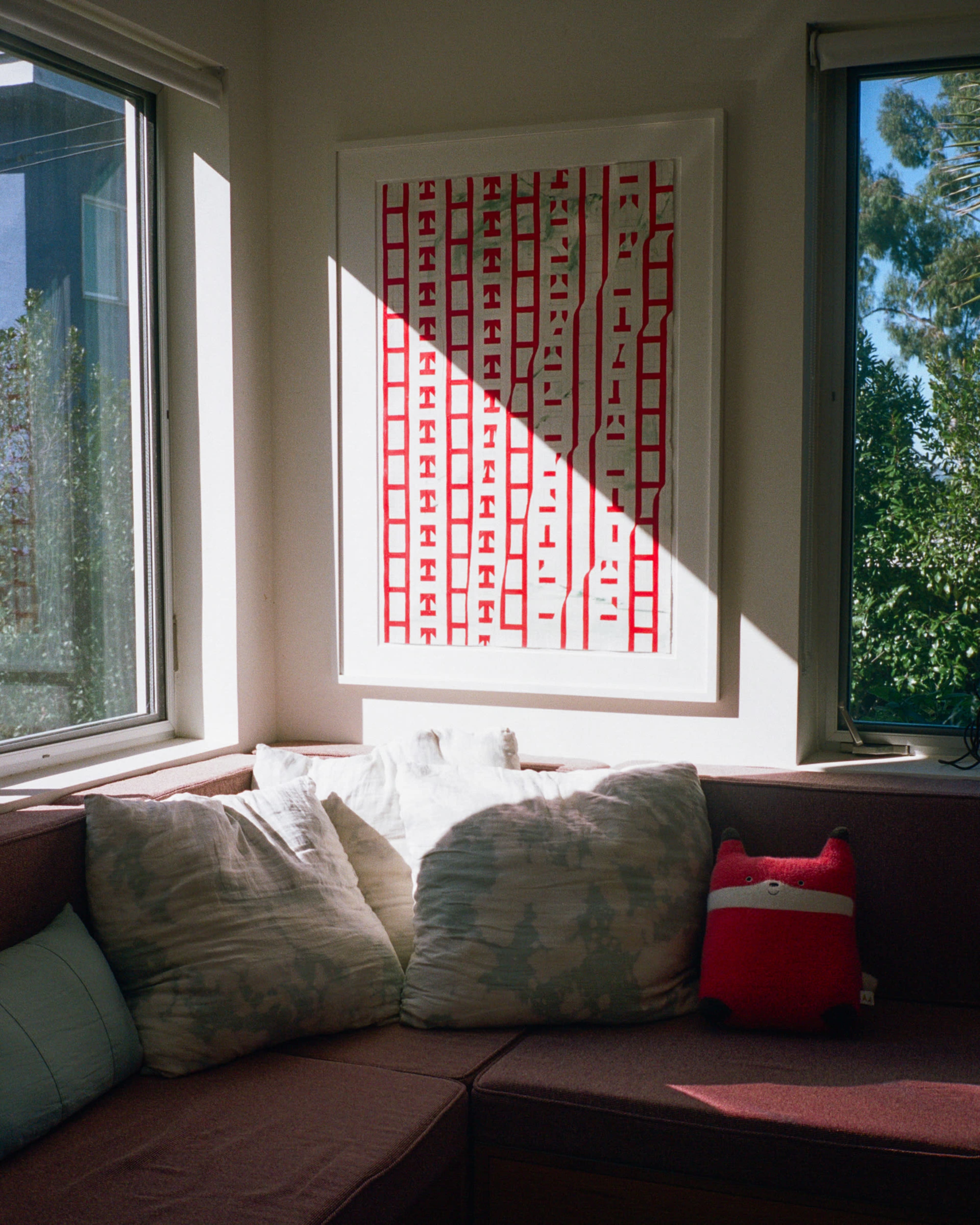 A couch with two large pillows sits in front of a window, and a framed artwork with red lines is mounted on the wall.