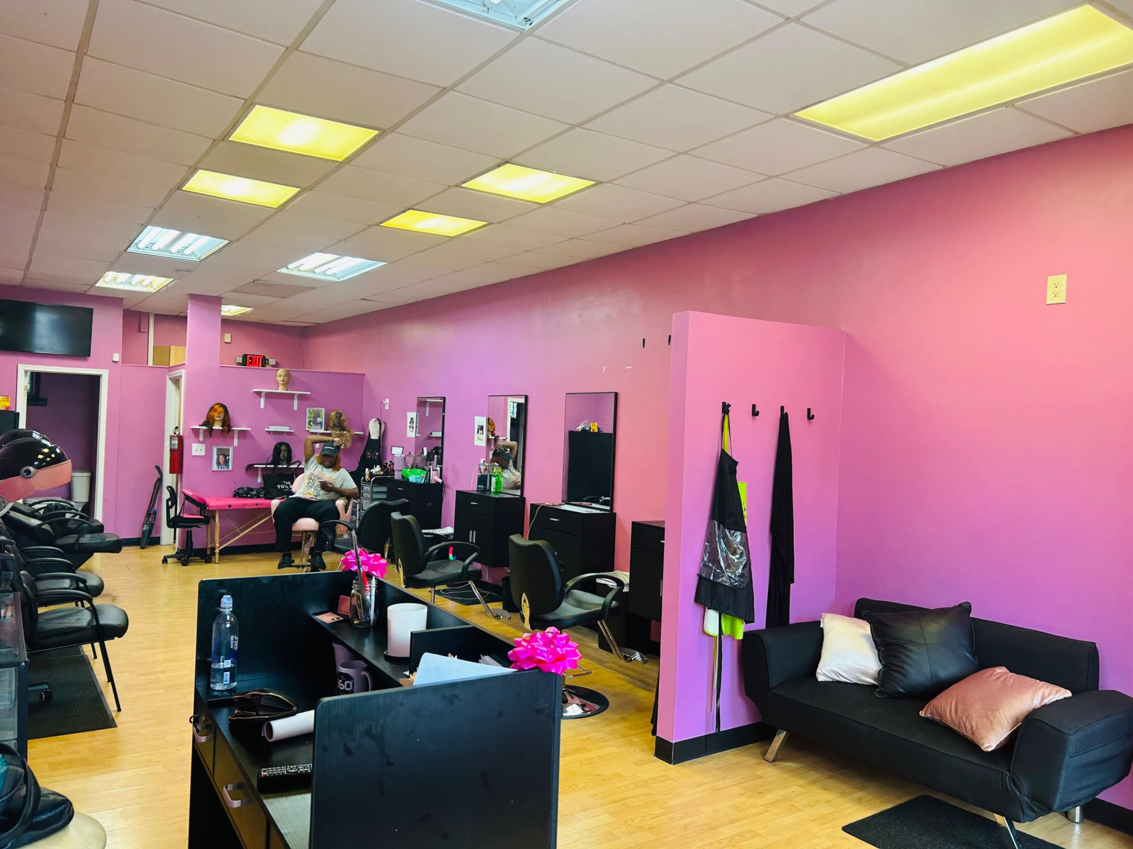 The image shows a hair salon interior with pink walls, black styling chairs, and a small waiting area featuring a dark sofa and decorative flowers.