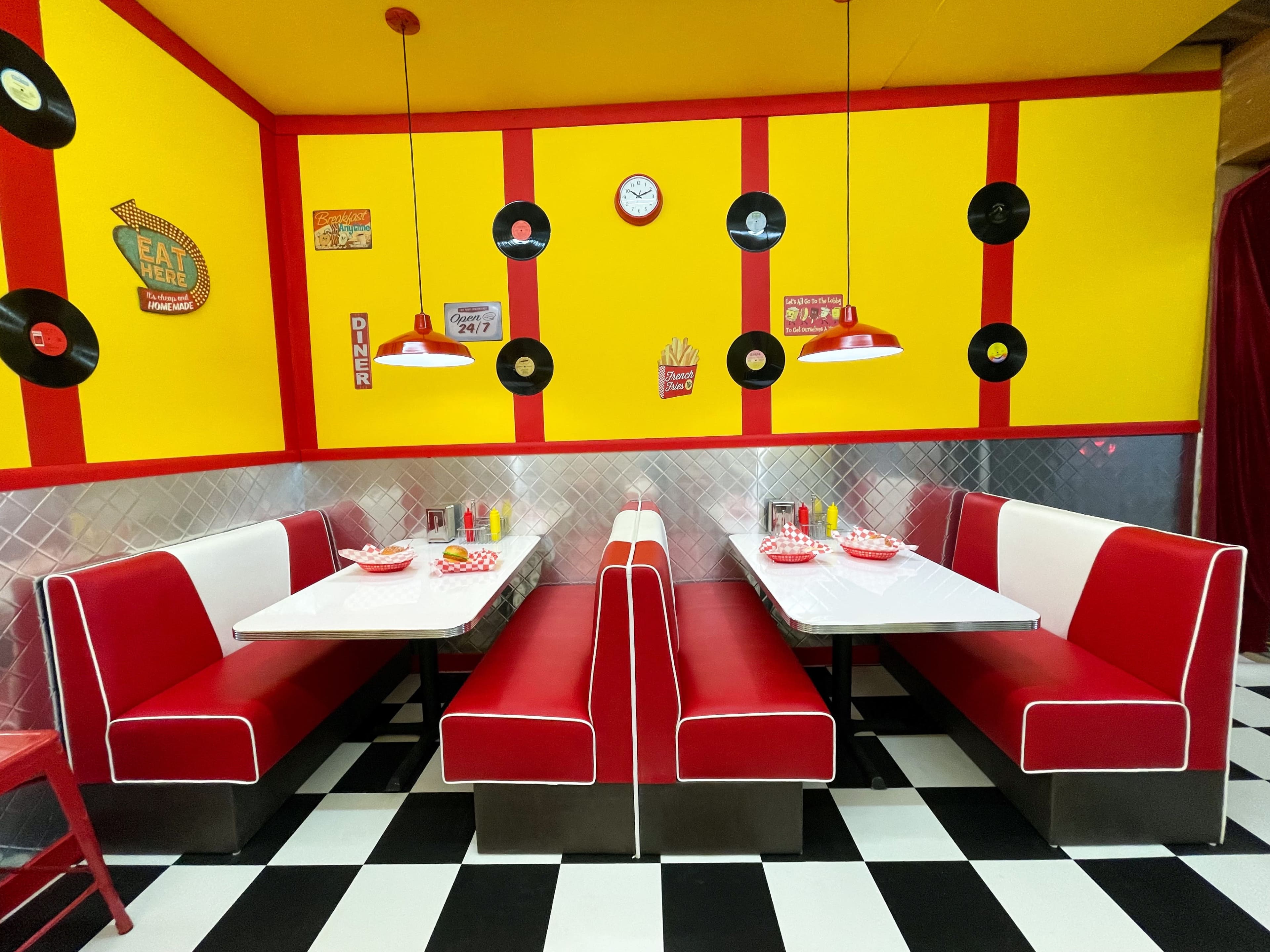 A bright diner interior with red and white booths, checkered flooring, and a yellow wall adorned with vintage posters and decorations.
