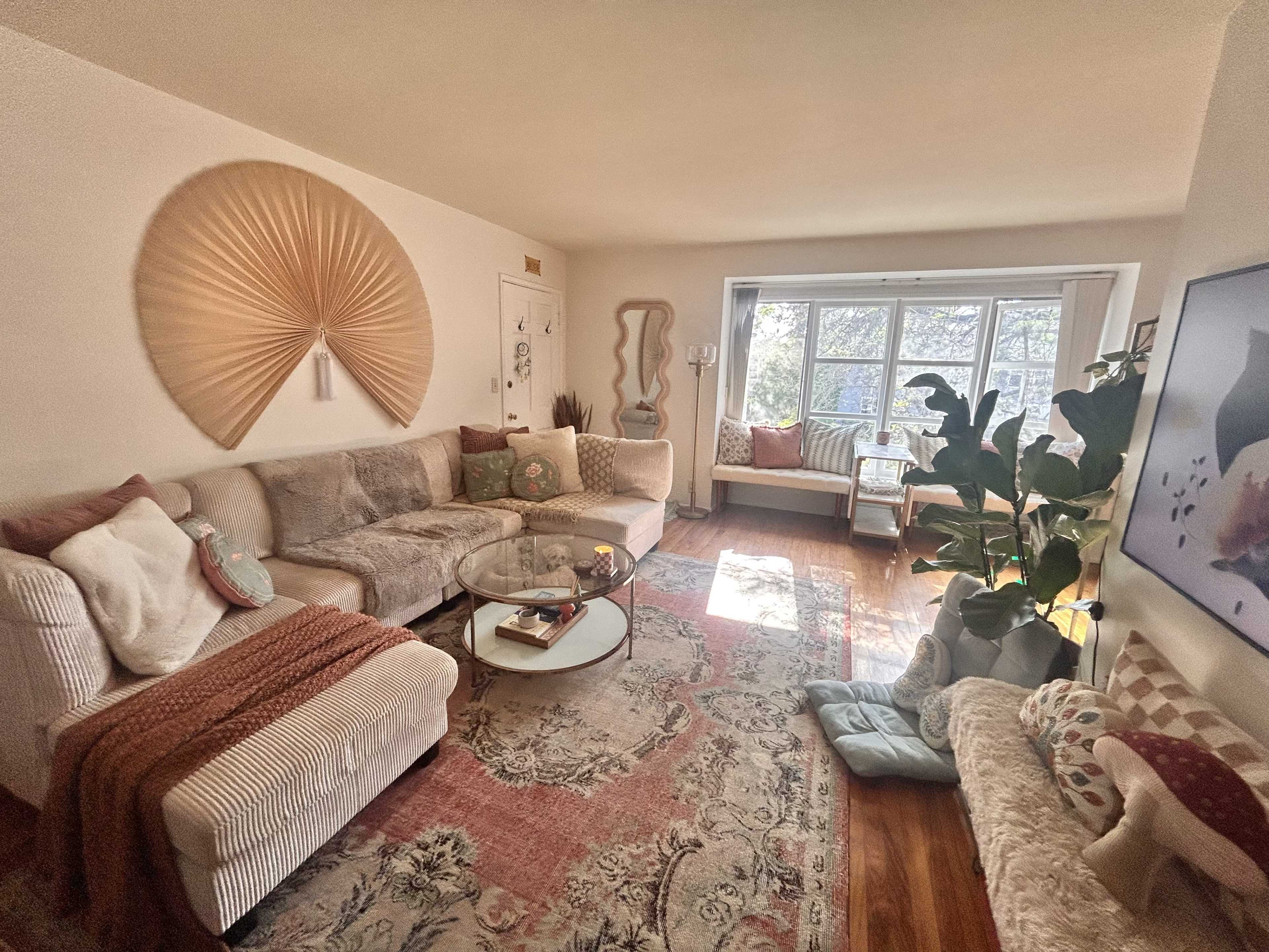 A living room features a large sectional sofa, a glass coffee table, a decorative wall panel, and a variety of plants beside a bright window.