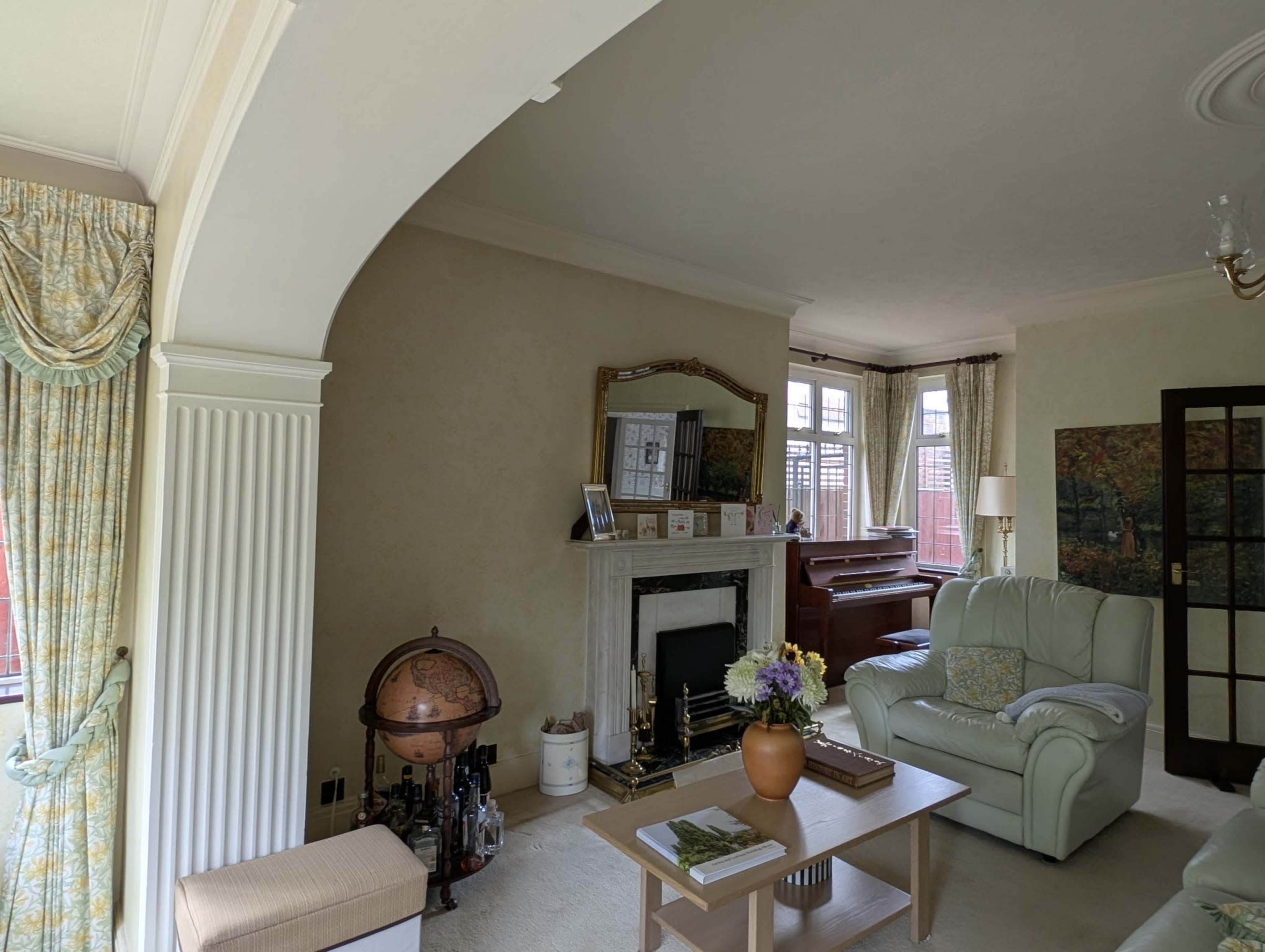 The image shows a living room with a fireplace, a piano, and light-colored furnishings, featuring large windows and a globe in the corner.