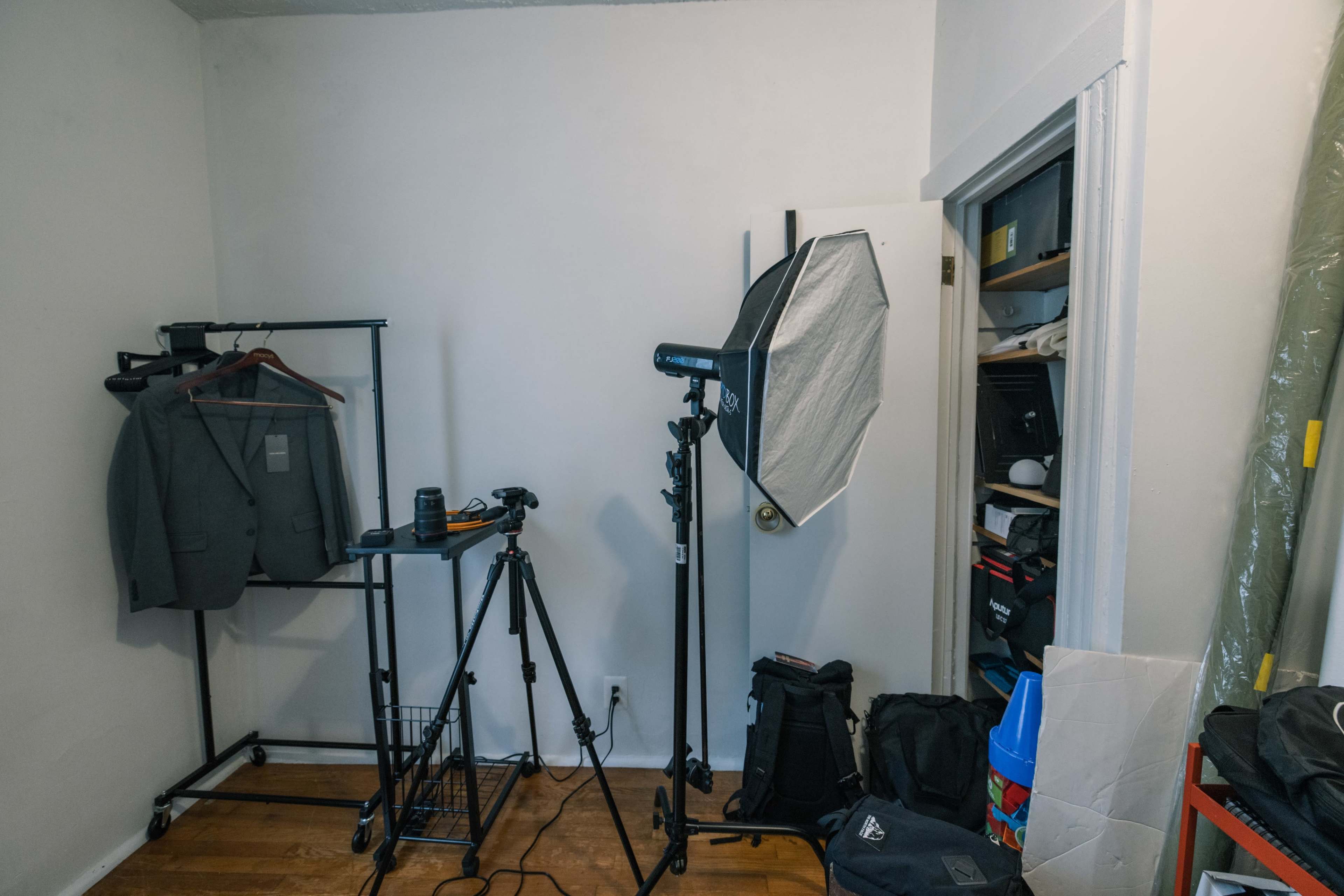A small photo studio setup with a clothing rack, camera equipment on a tripod, and a lighting umbrella in a cramped room.