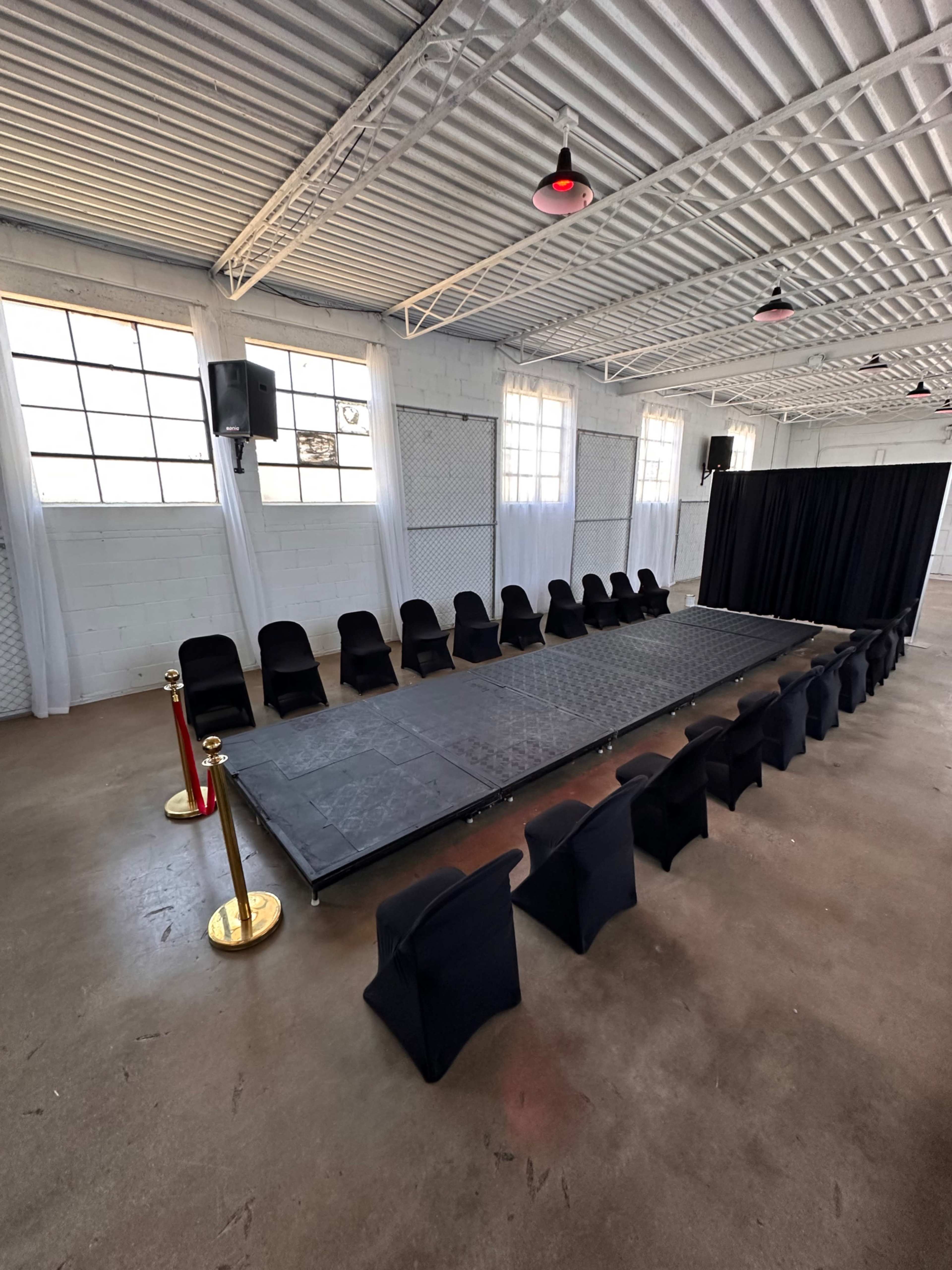 The image shows a runway set for a fashion show, featuring a long black platform with chairs arranged on either side and a black curtain at one end.