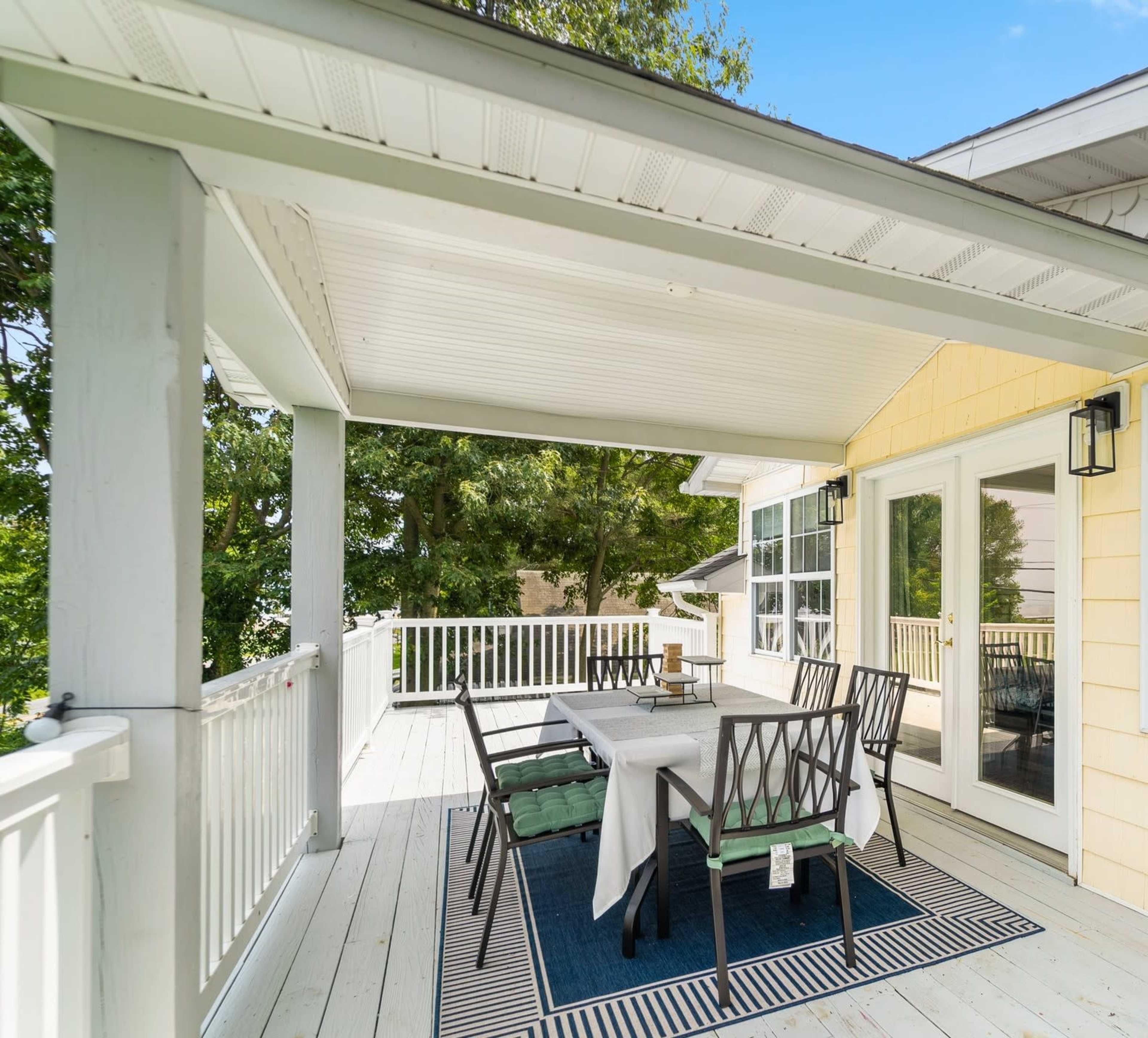 A patio features a dining table surrounded by chairs, situated under a covered porch with access to a house.