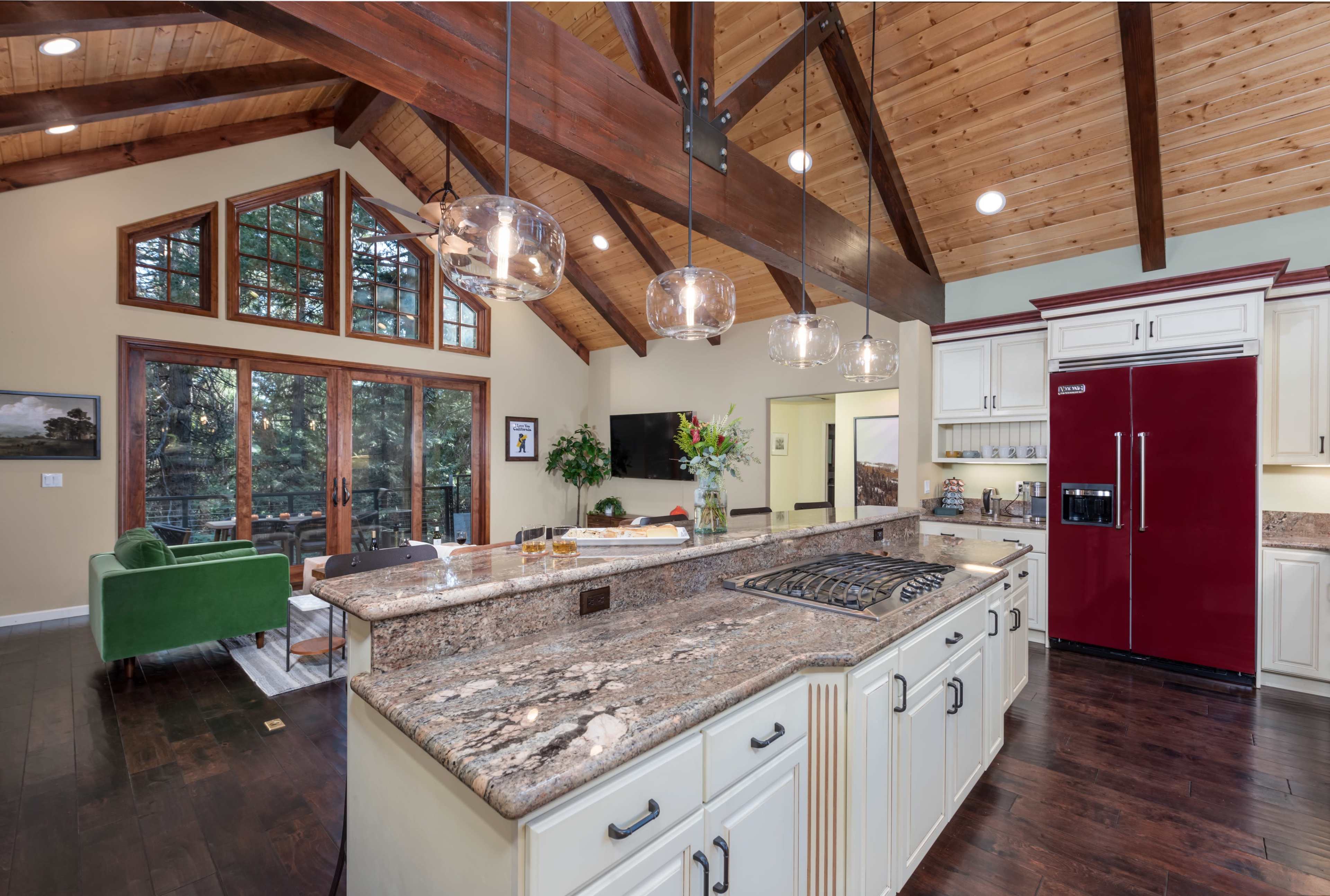 A spacious kitchen features a large granite island, modern appliances, and vaulted wooden beams, with large windows providing views of the outdoors.