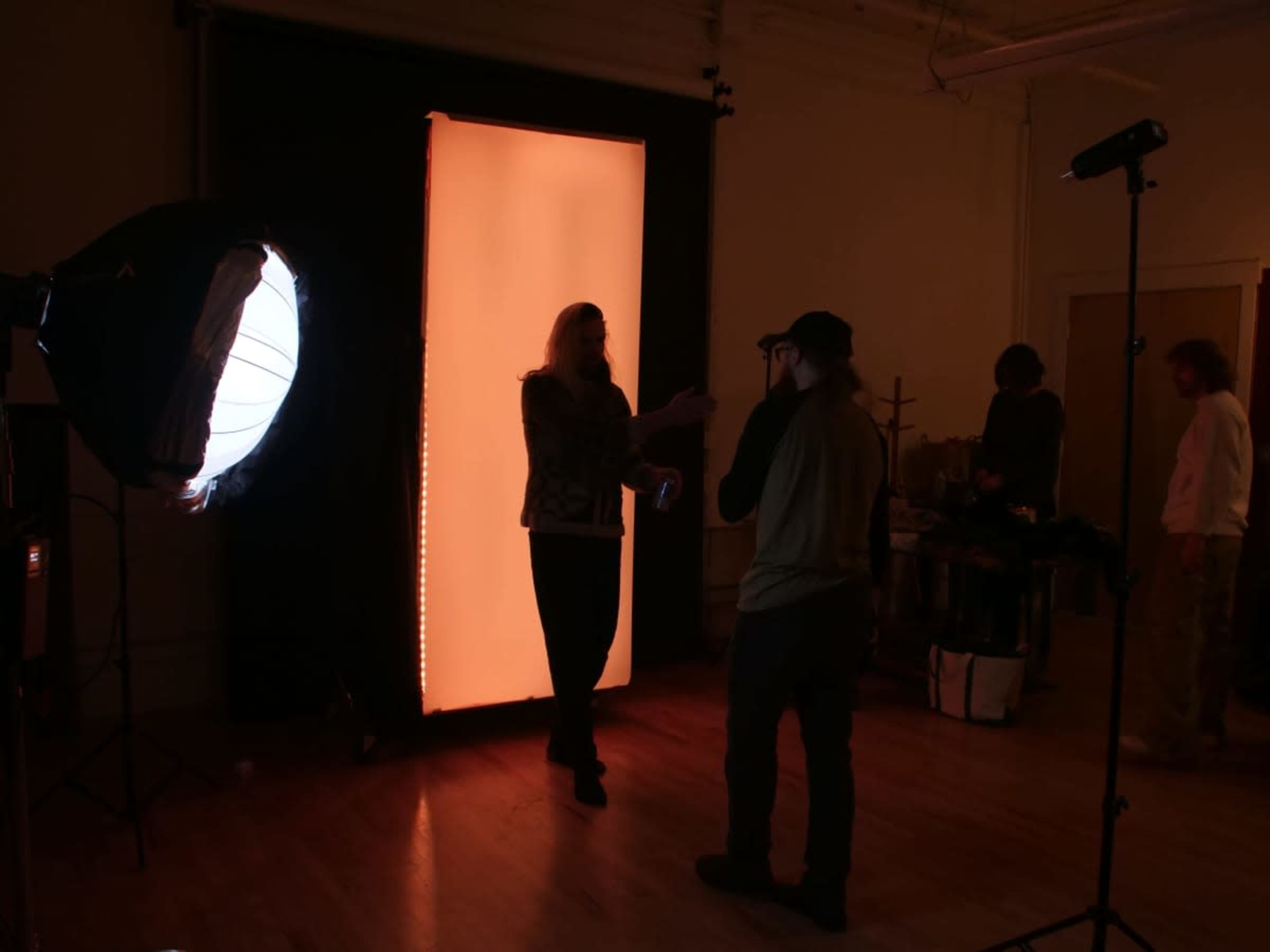 A person with long hair interacts with another individual in a photography studio illuminated by an orange backdrop and lighting equipment.