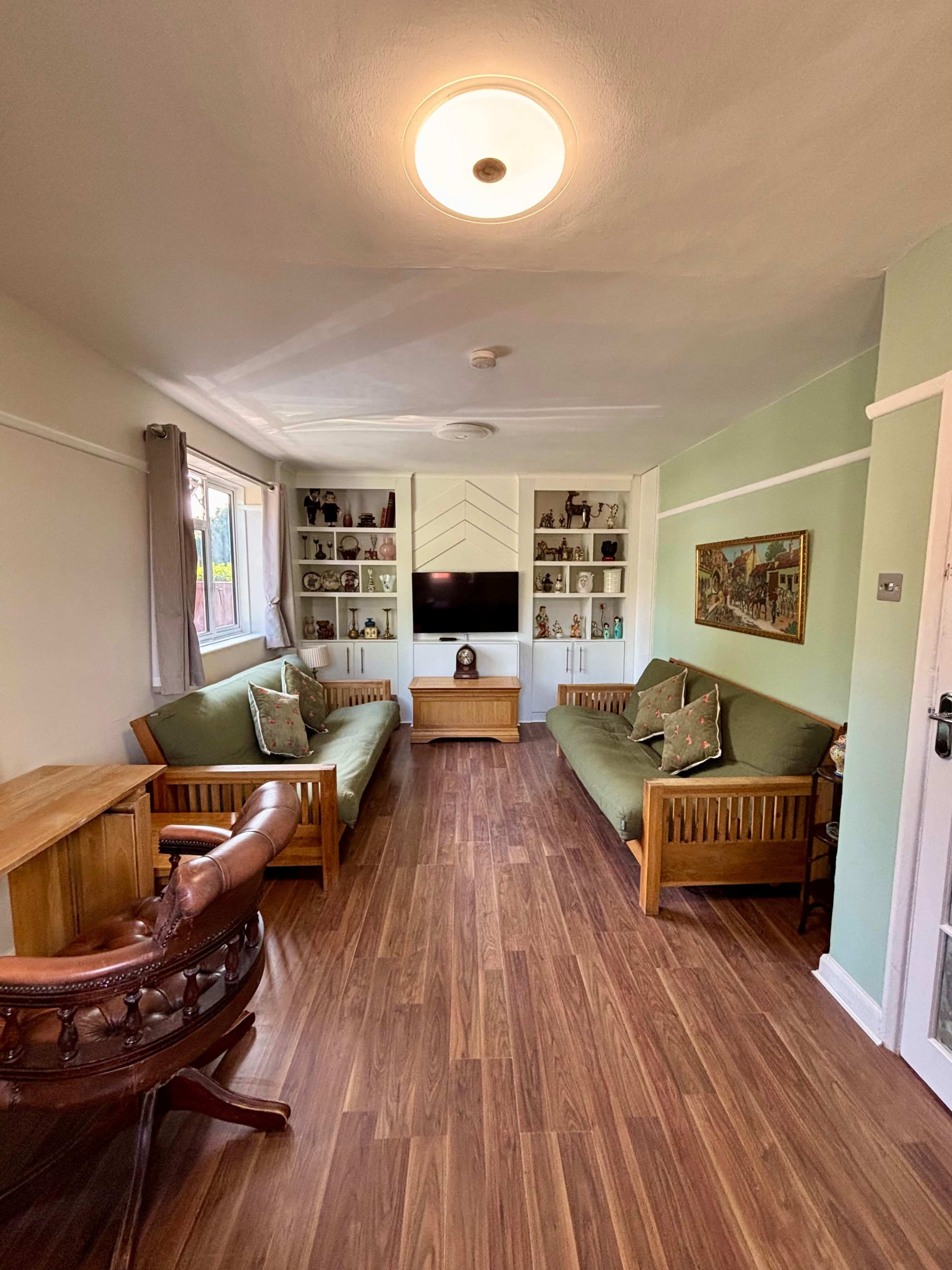 The image shows a living room with two wooden sofa sets, a television mounted on the wall, and shelves displaying decorative items.