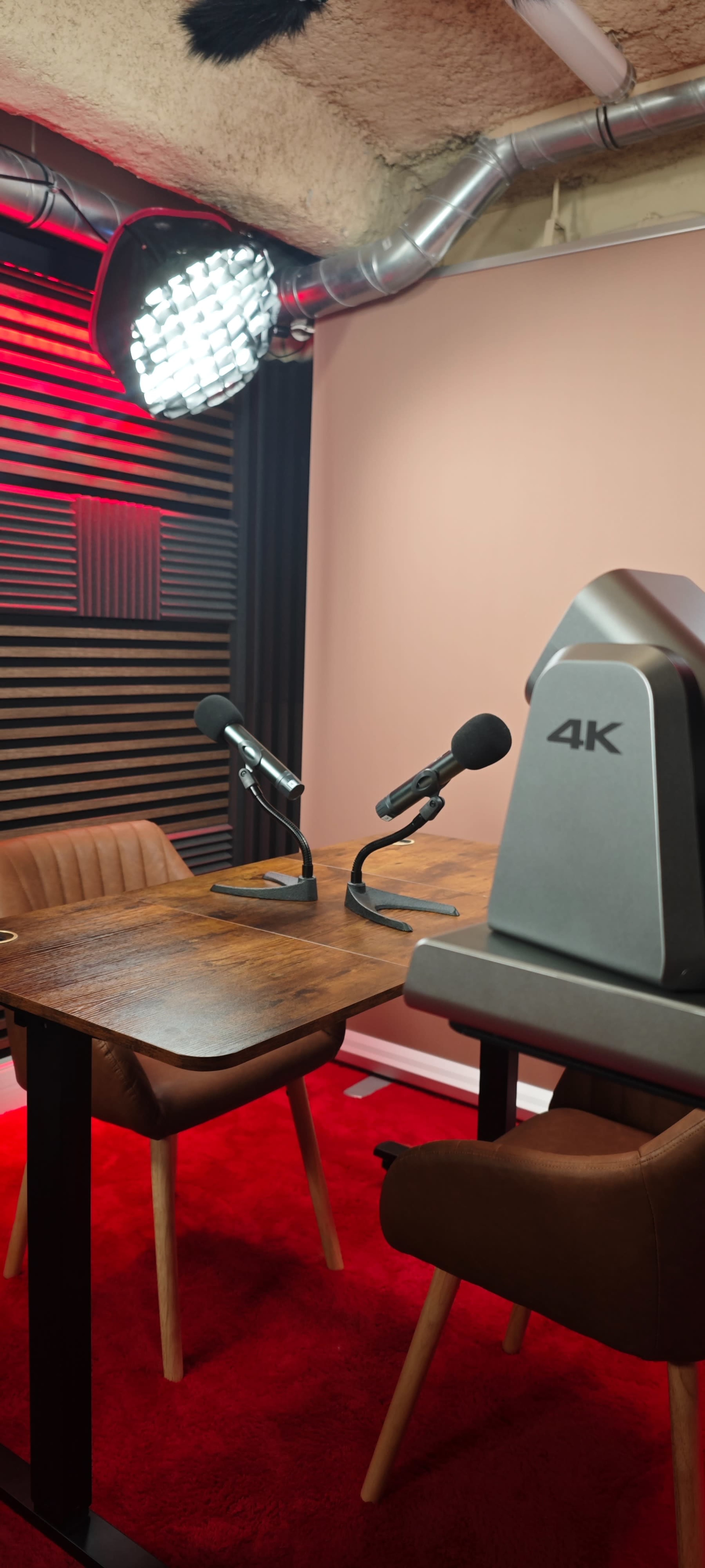 The image shows a podcast recording setup with two microphones positioned at a wooden table in a studio featuring red and black decor.