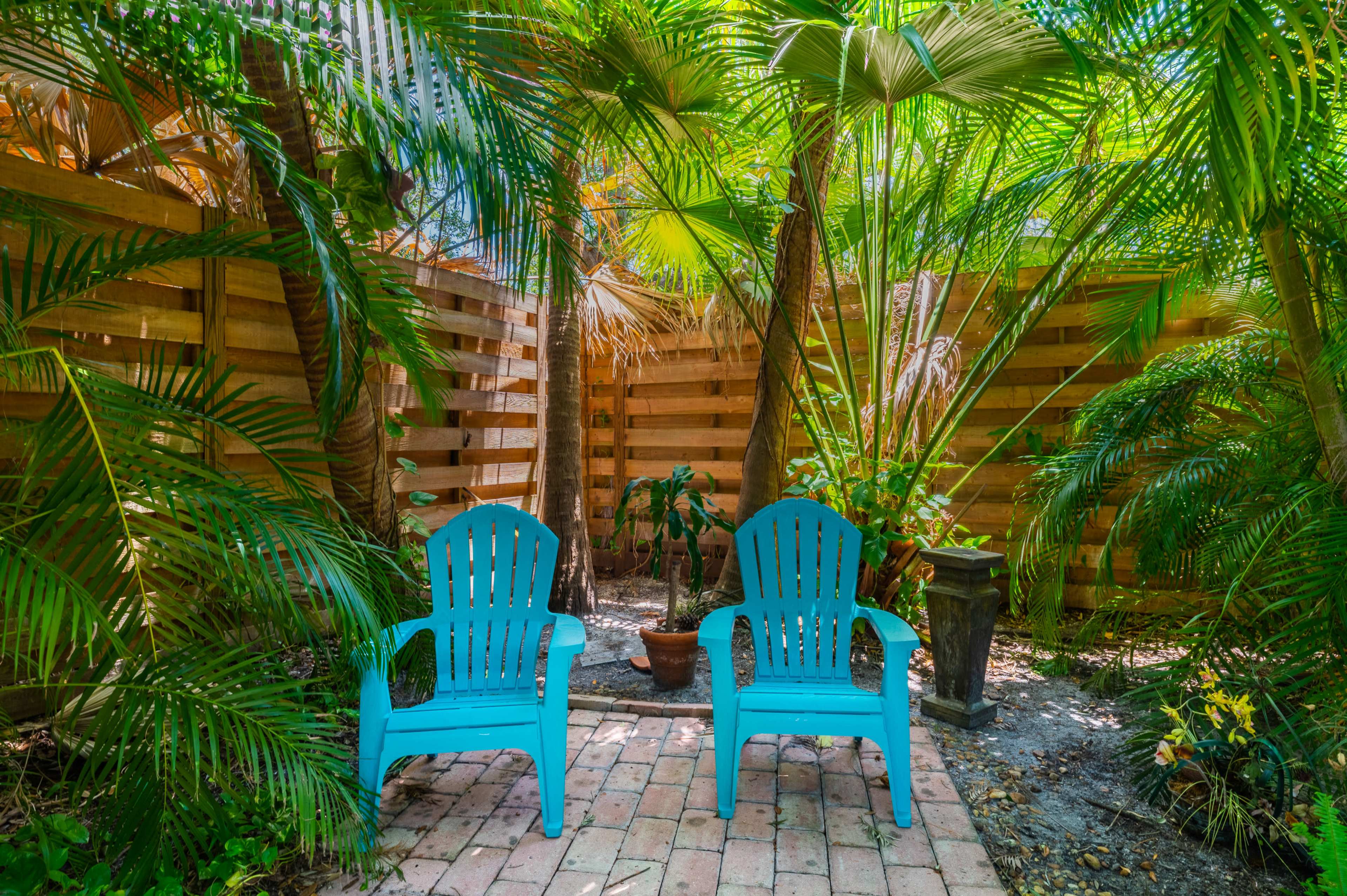 Two teal adirondack chairs are positioned on a brick patio surrounded by lush tropical plants and wooden fencing.