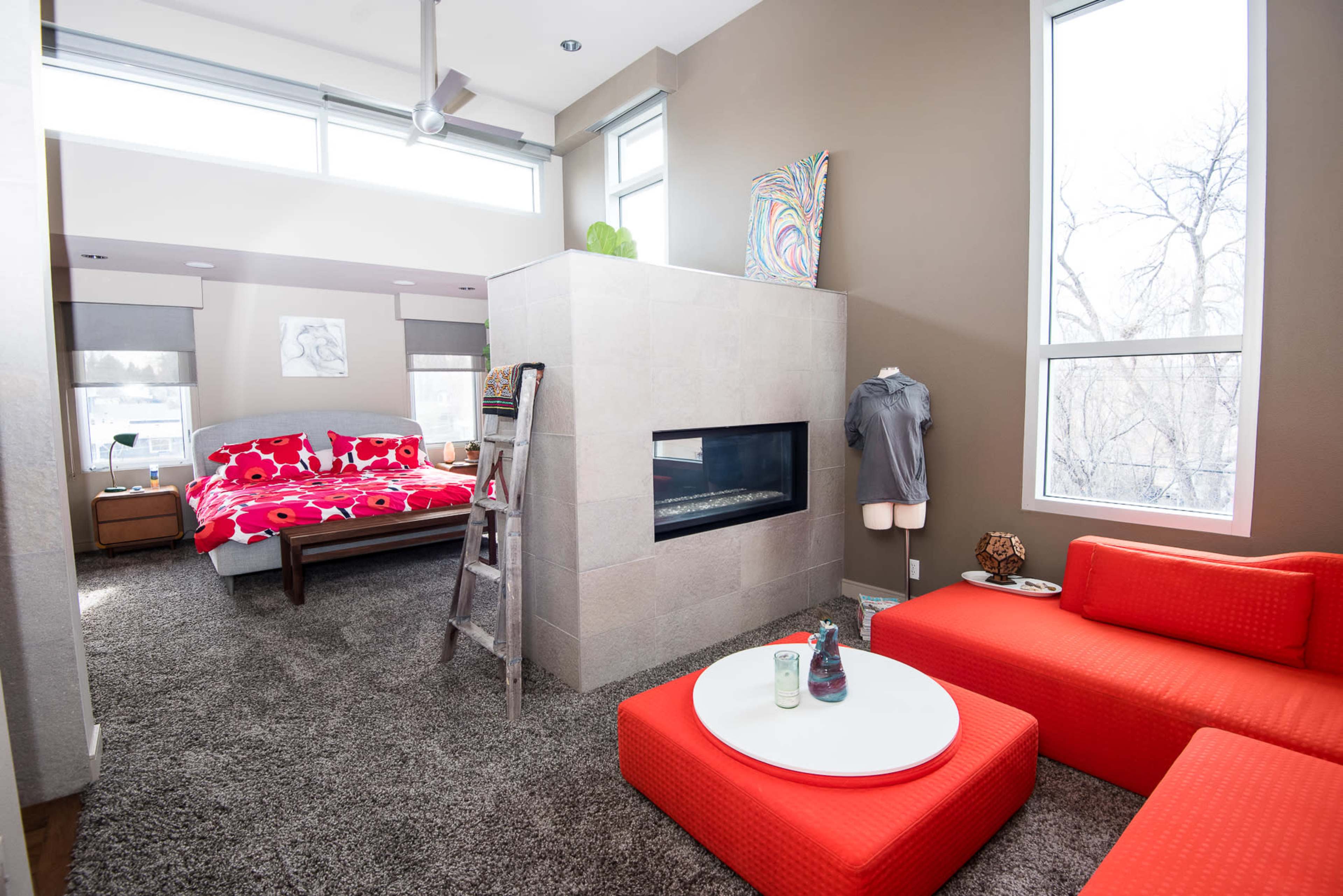 The room features a modern design with a bedroom area containing a red floral bedspread, a sitting area with orange furniture, and a fireplace as a central element.