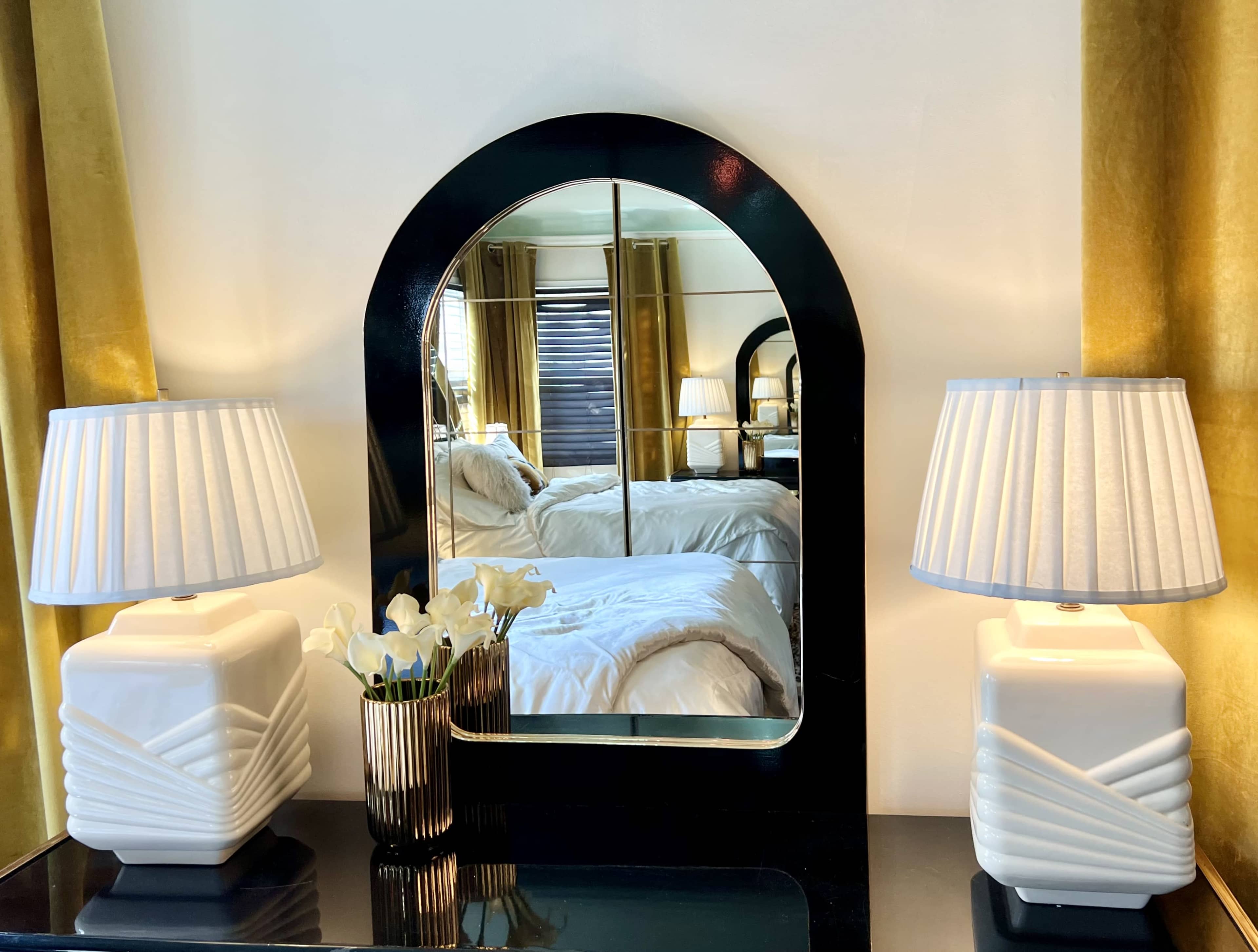 A black-framed mirror flanked by two white lamps on a dark surface, with a vase of flowers and a bed visible in the reflection.