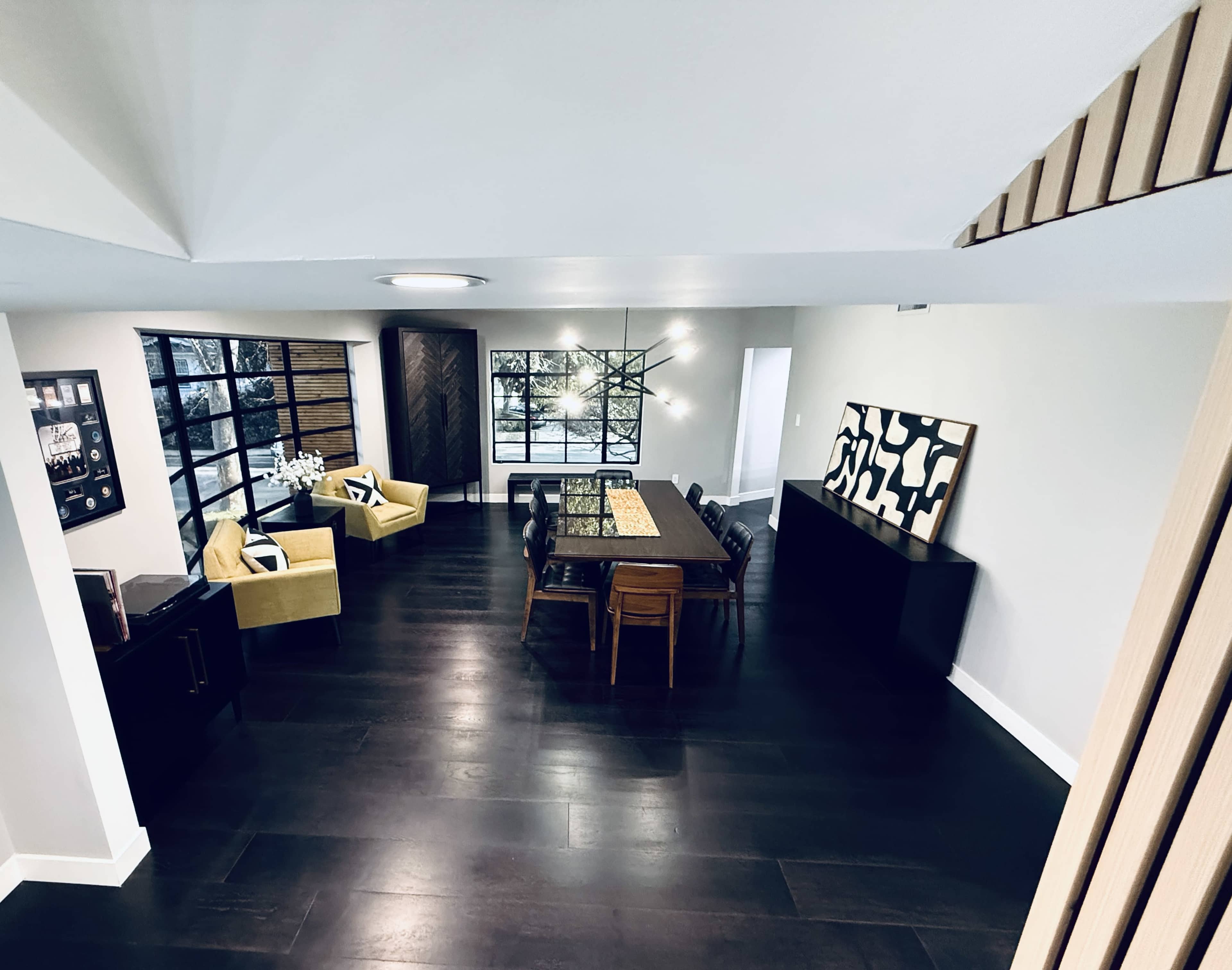 The image shows a spacious, modern dining area featuring a large table, wooden chairs, and contemporary decor, with windows providing natural light and a view of outdoor greenery.