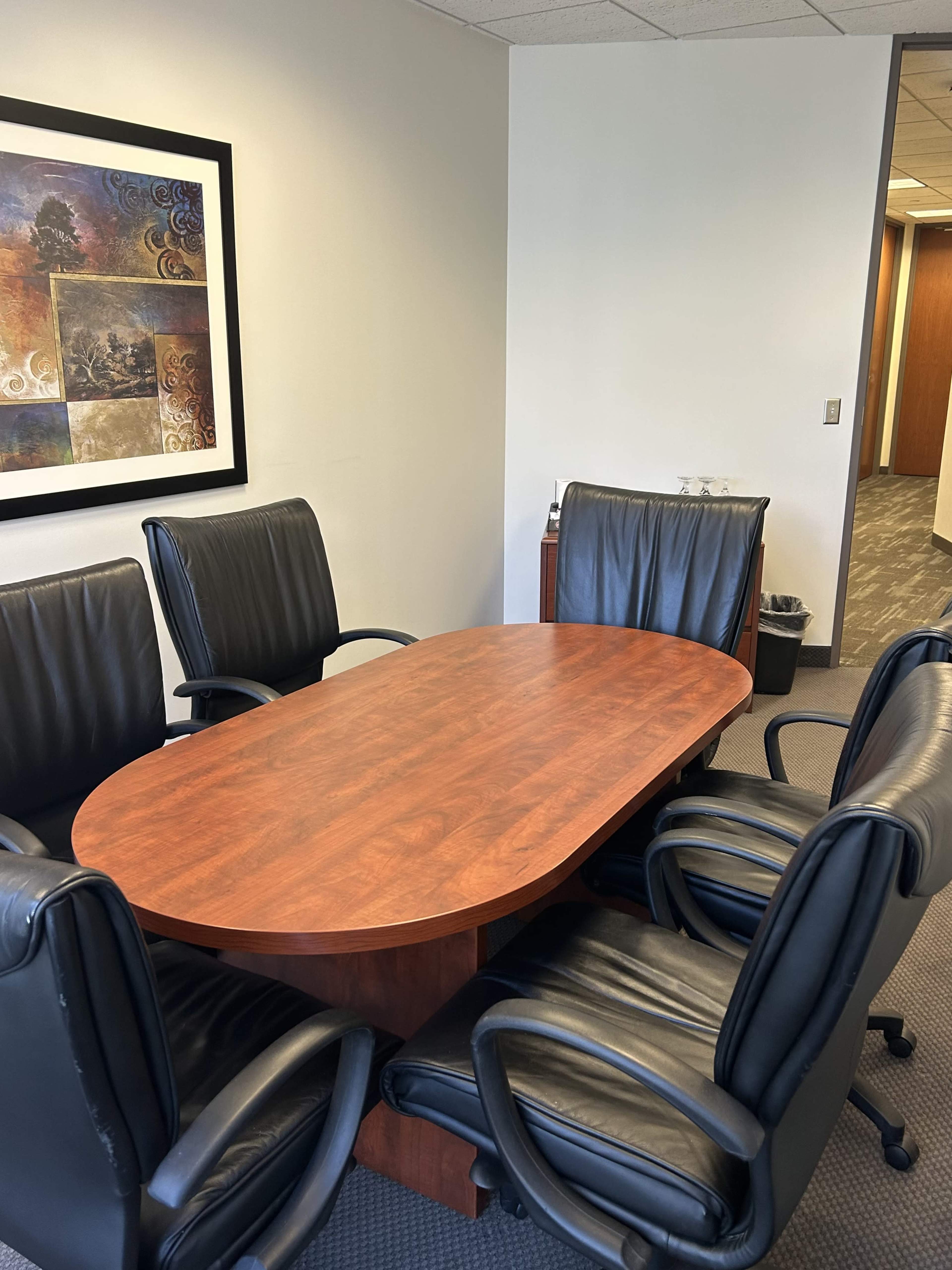 The image shows a conference room with a wooden oval table surrounded by five black leather chairs, along with a framed artwork on the wall.