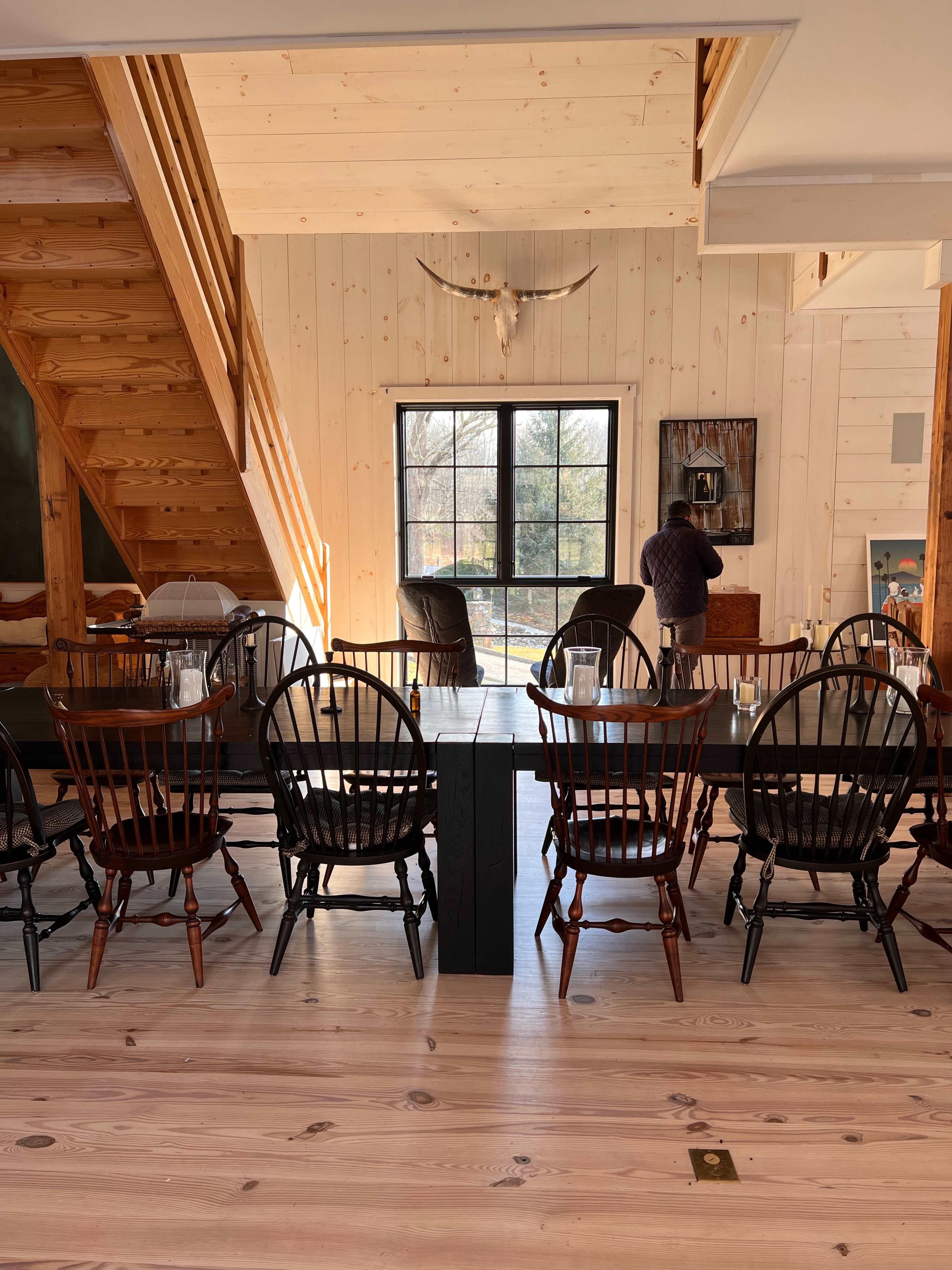 A spacious dining area features a long black table with six wooden chairs, situated beside a stairway and a large window that overlooks a scenic outdoor view.