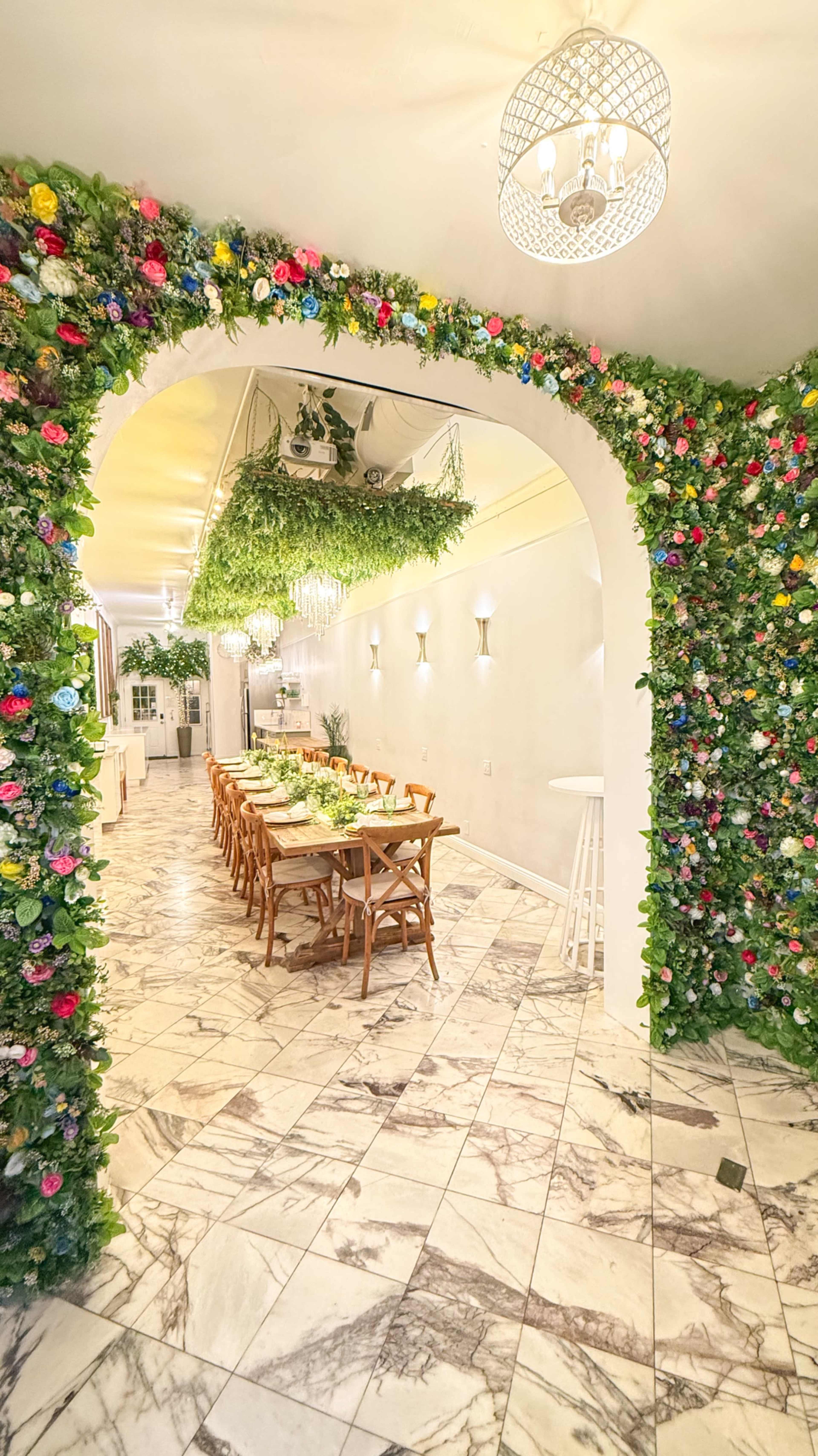 A bright restaurant interior features a long dining table surrounded by floral and greenery decorations along the walls and ceiling.